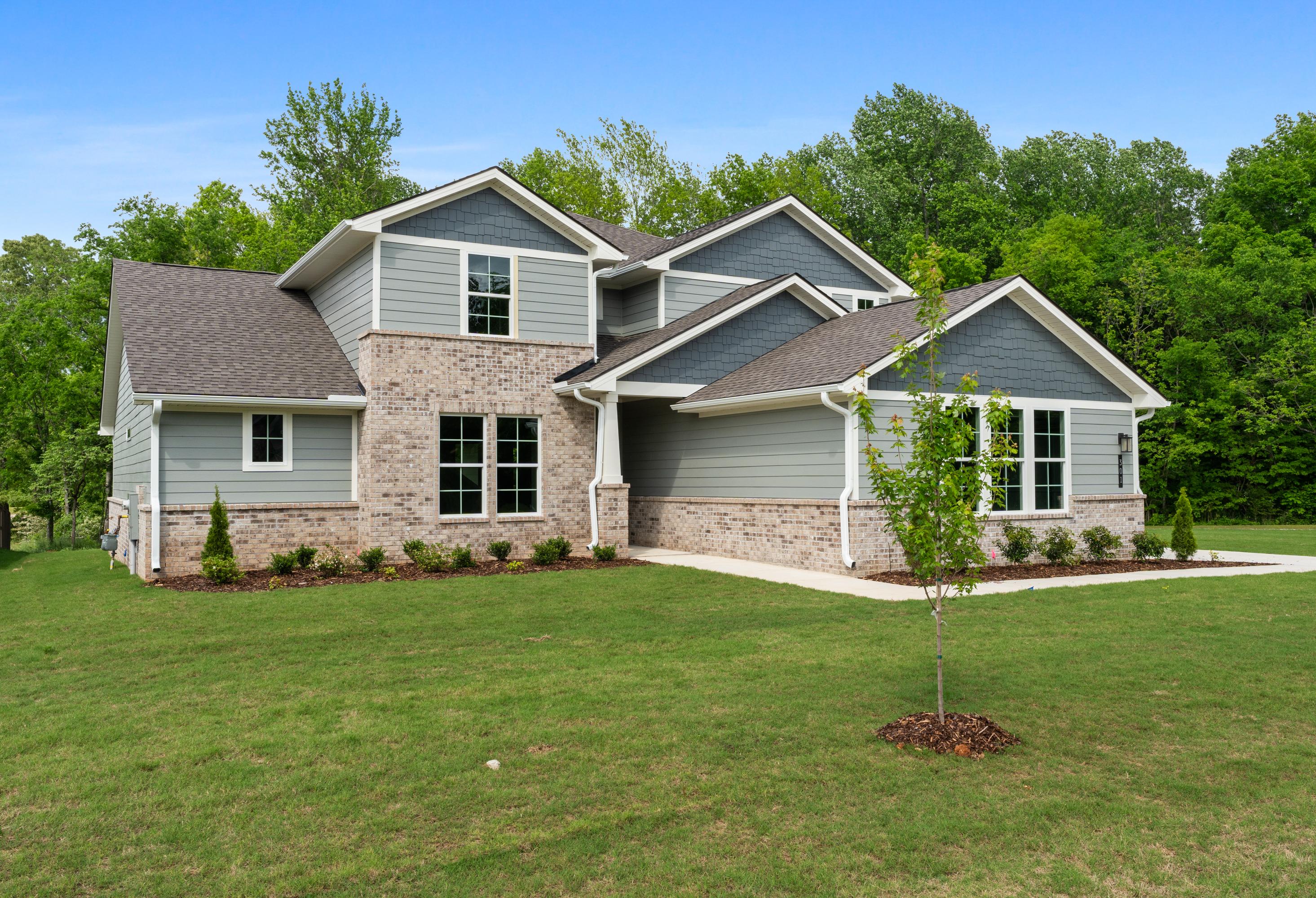 Two-story Avalon D home elevation with gray siding, brick accents, shingled roof, and lush green landscaping