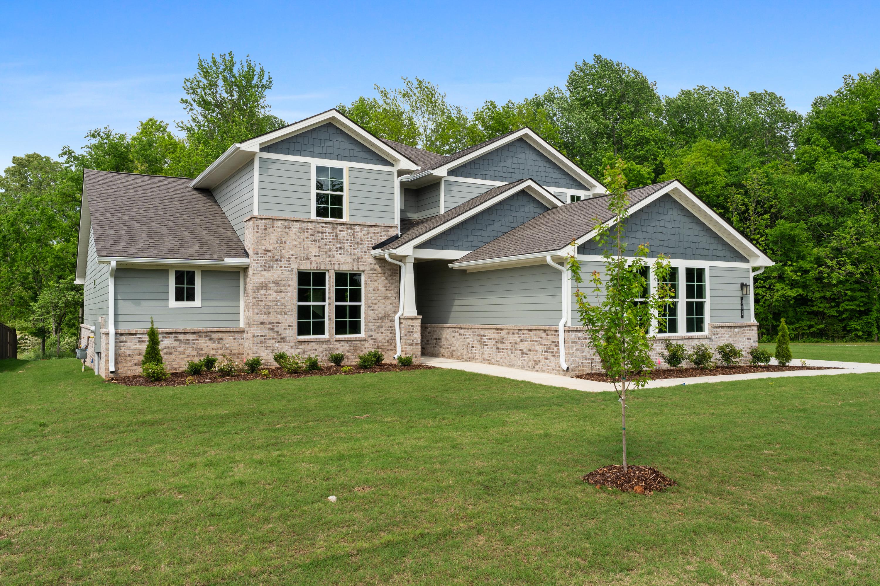 Two-story Avalon D home elevation with gray siding, brick accents, shingled roof, and lush green landscaping