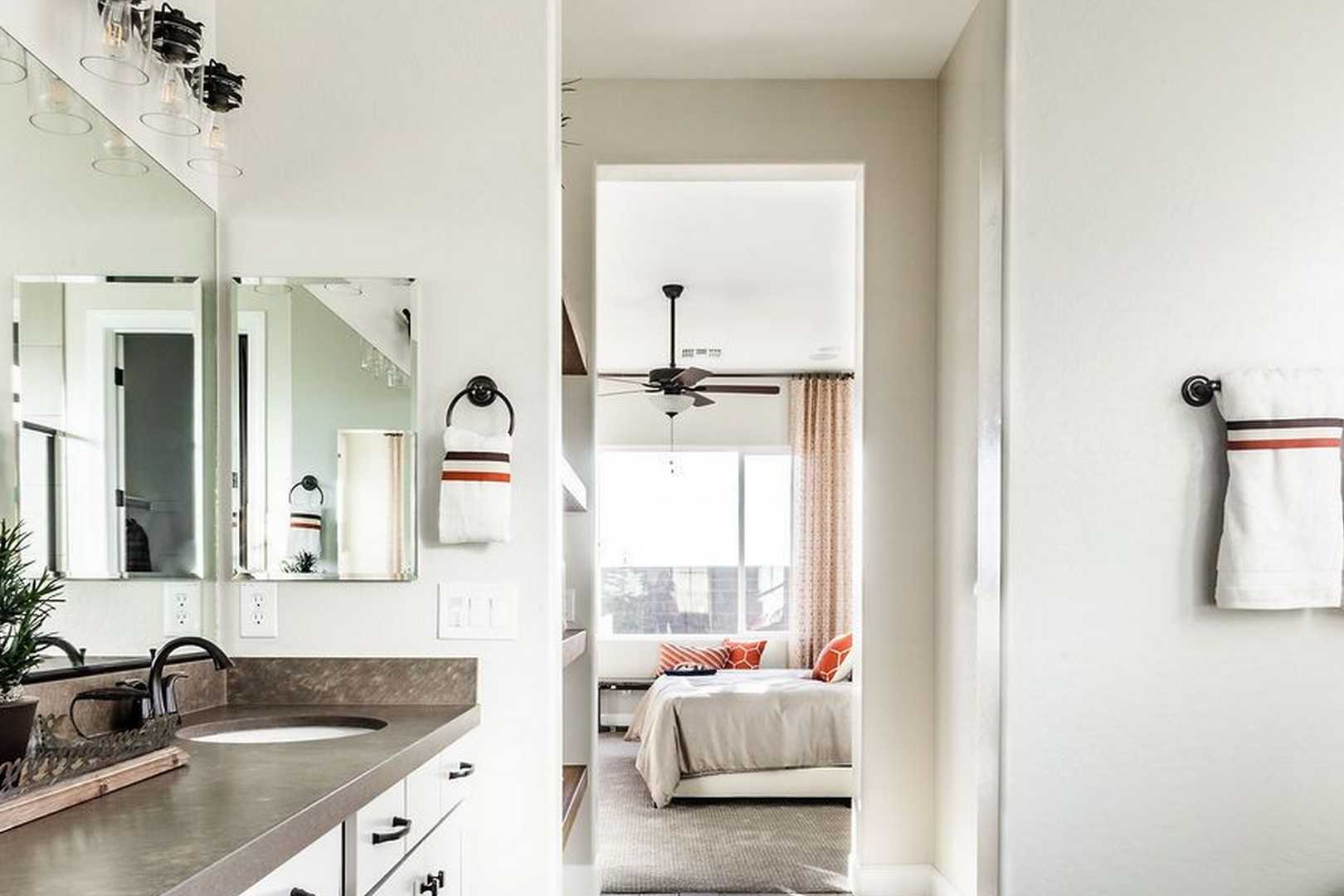 Spacious master bathroom in The Monarch home featuring double vanity, mirrors, towels, plants, and adjacent bedroom with ceiling fan