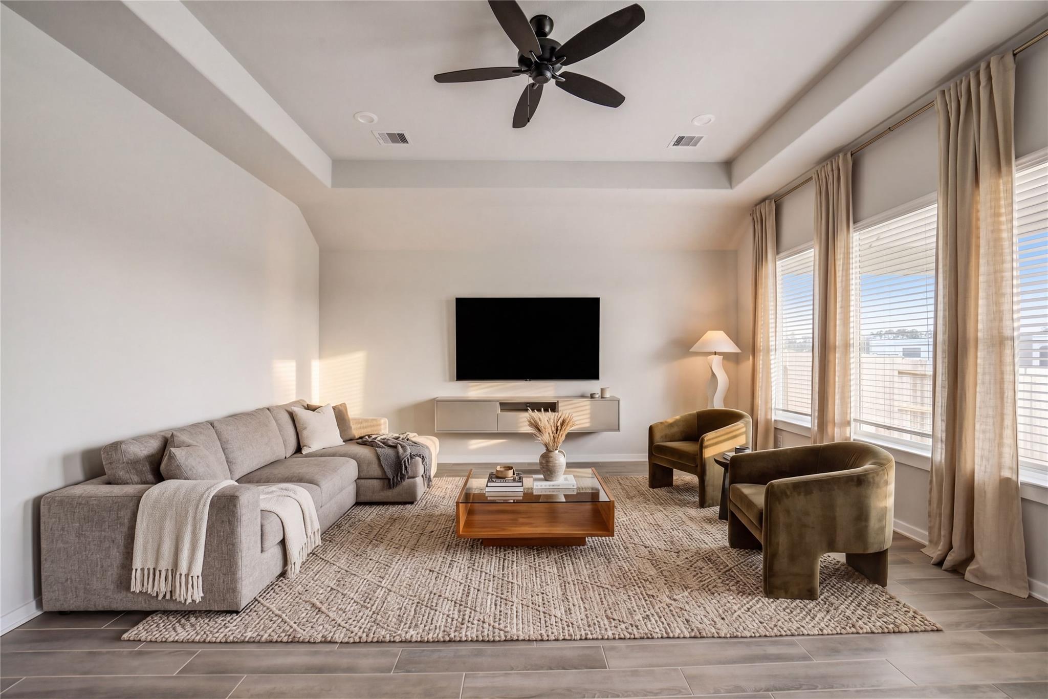 Modern living room with gray sectional sofa, wooden coffee table, green velvet chairs, mounted TV, ceiling fan, and large windows in Davidson Homes The Acadia C, Windmill Estates Magnolia TX