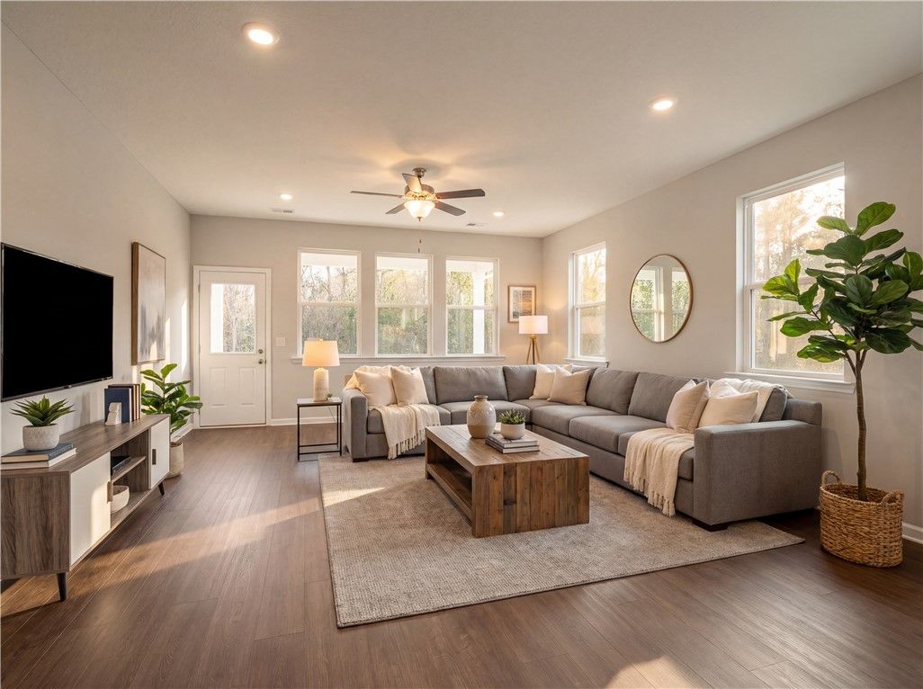 Cozy living room featuring gray L-shaped sectional, wooden coffee table, TV console, and large windows in Evermore Homes The Orion, Cusseta, Alabama