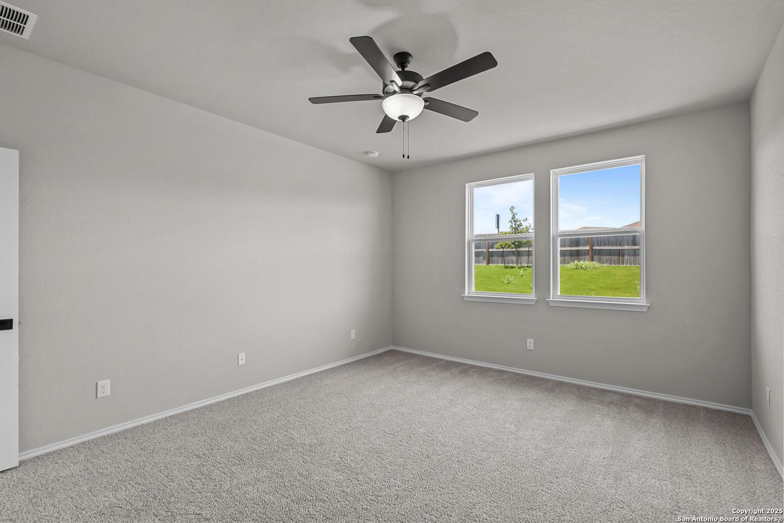 Bright bedroom with gray walls, ceiling fan, carpet floor, and windows overlooking green lawn in Davidson Homes The Daphne K, San Antonio