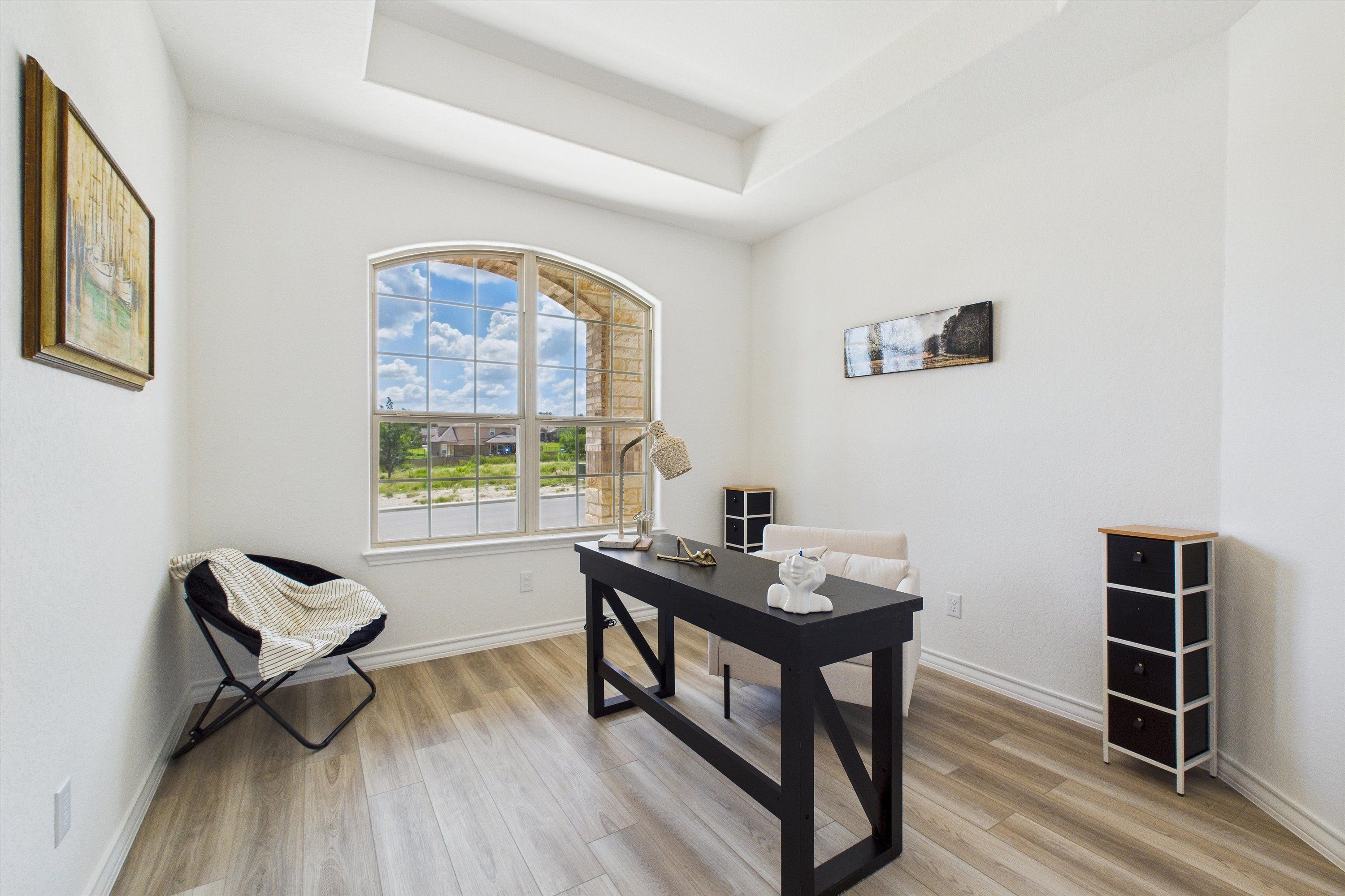Bright home office with arched window overlooking lush green field, farmhouse desk, armchair, and white cabinet in Summerlin C, Castroville, Texas