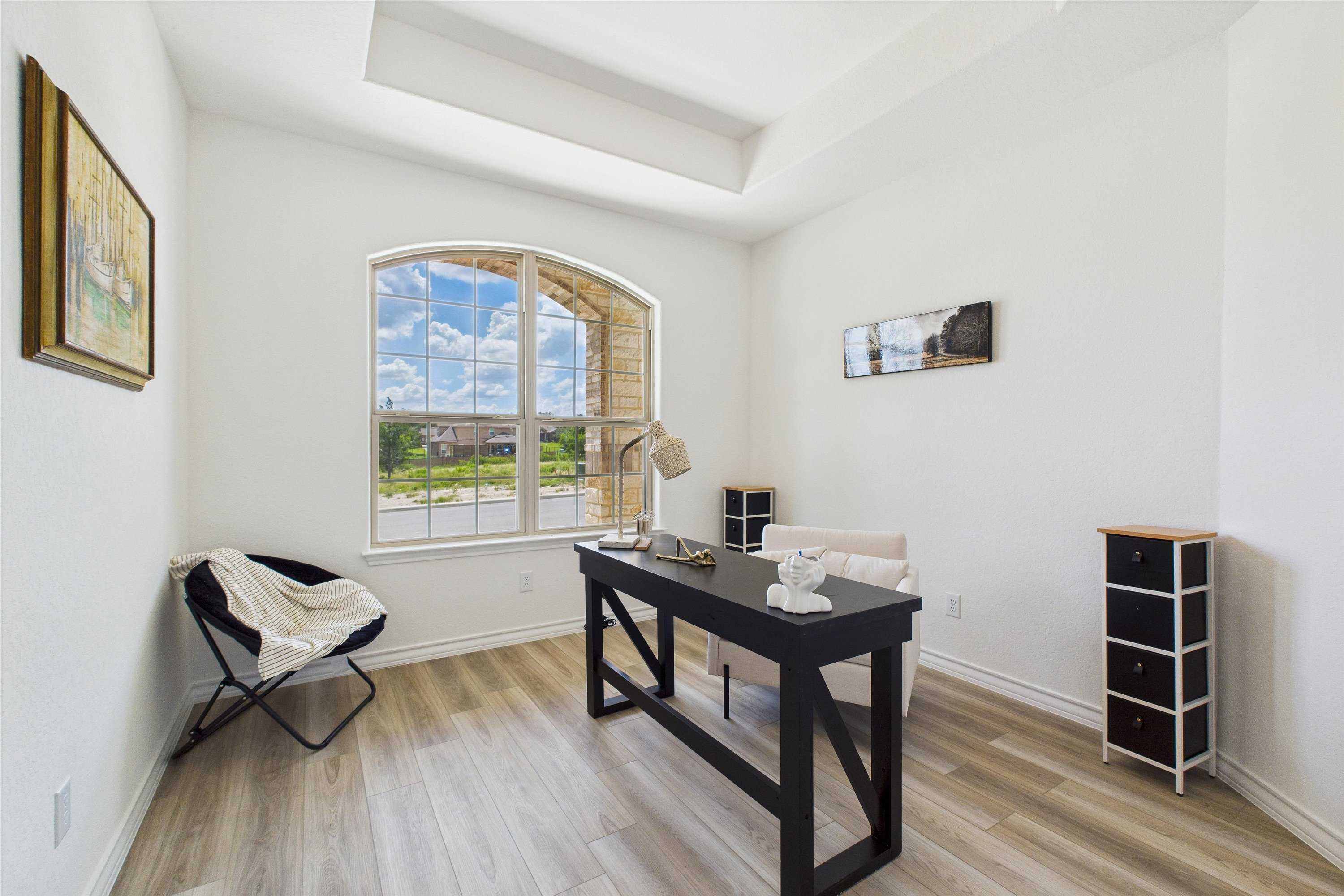 Bright home office with arched window overlooking lush green field, farmhouse desk, armchair, and white cabinet in Summerlin C, Castroville, Texas
