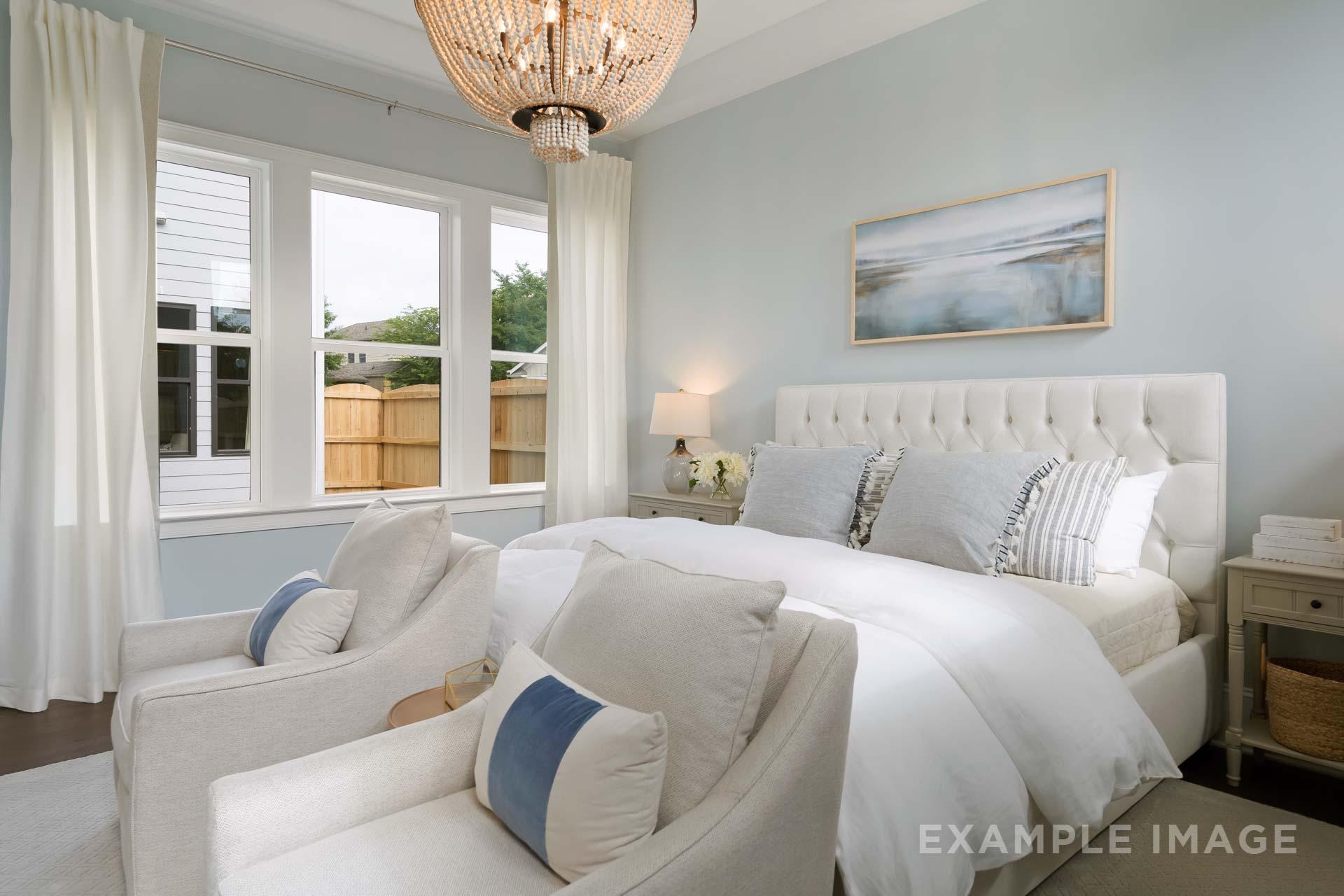 Spacious master suite in The Seaside C featuring white tufted bed, armchairs, rattan chandelier, and seascape art on light blue walls