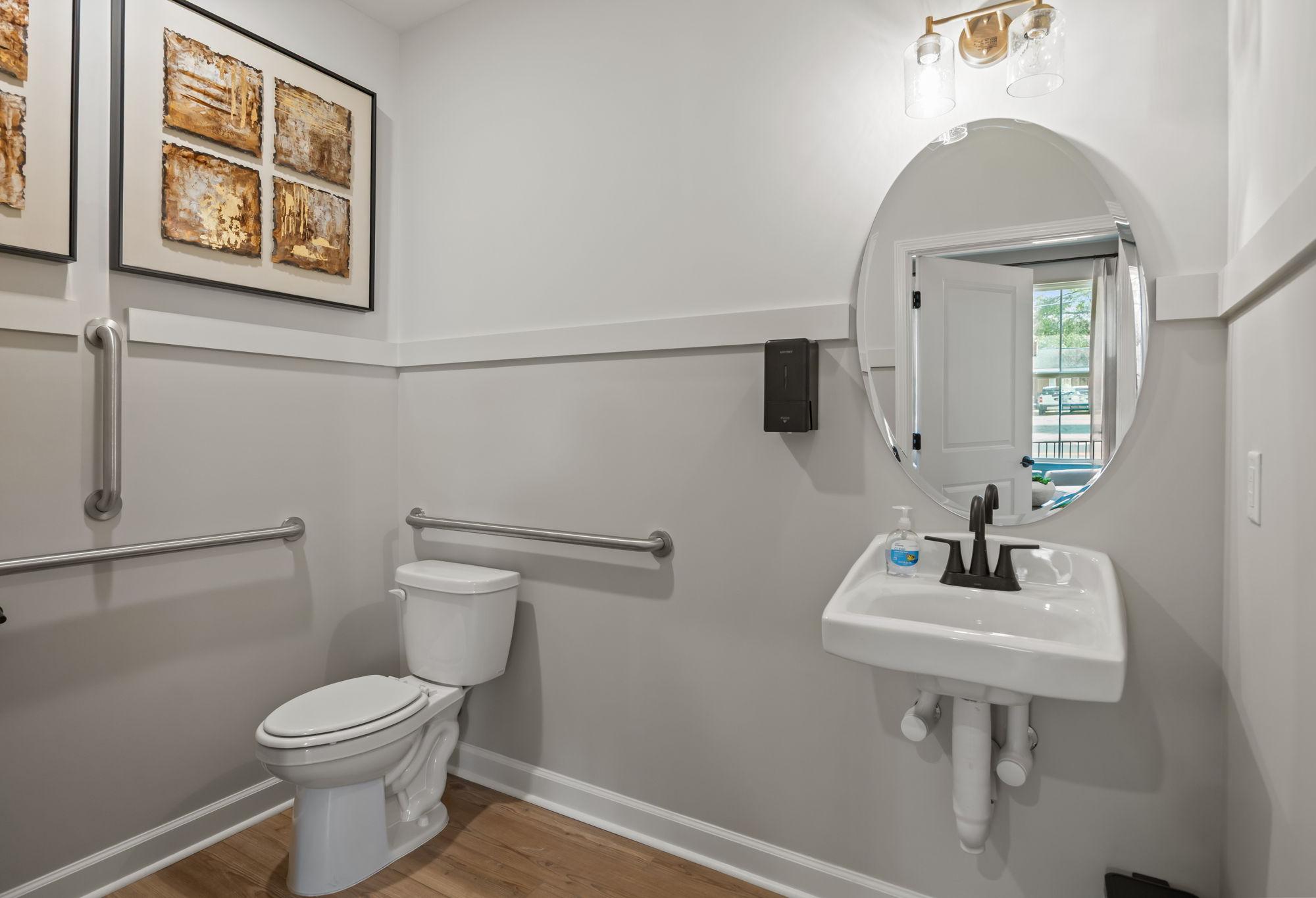 Accessible powder room in The Mitchell interior, featuring a wall-mounted sink, oval mirror, and safety grab bars.