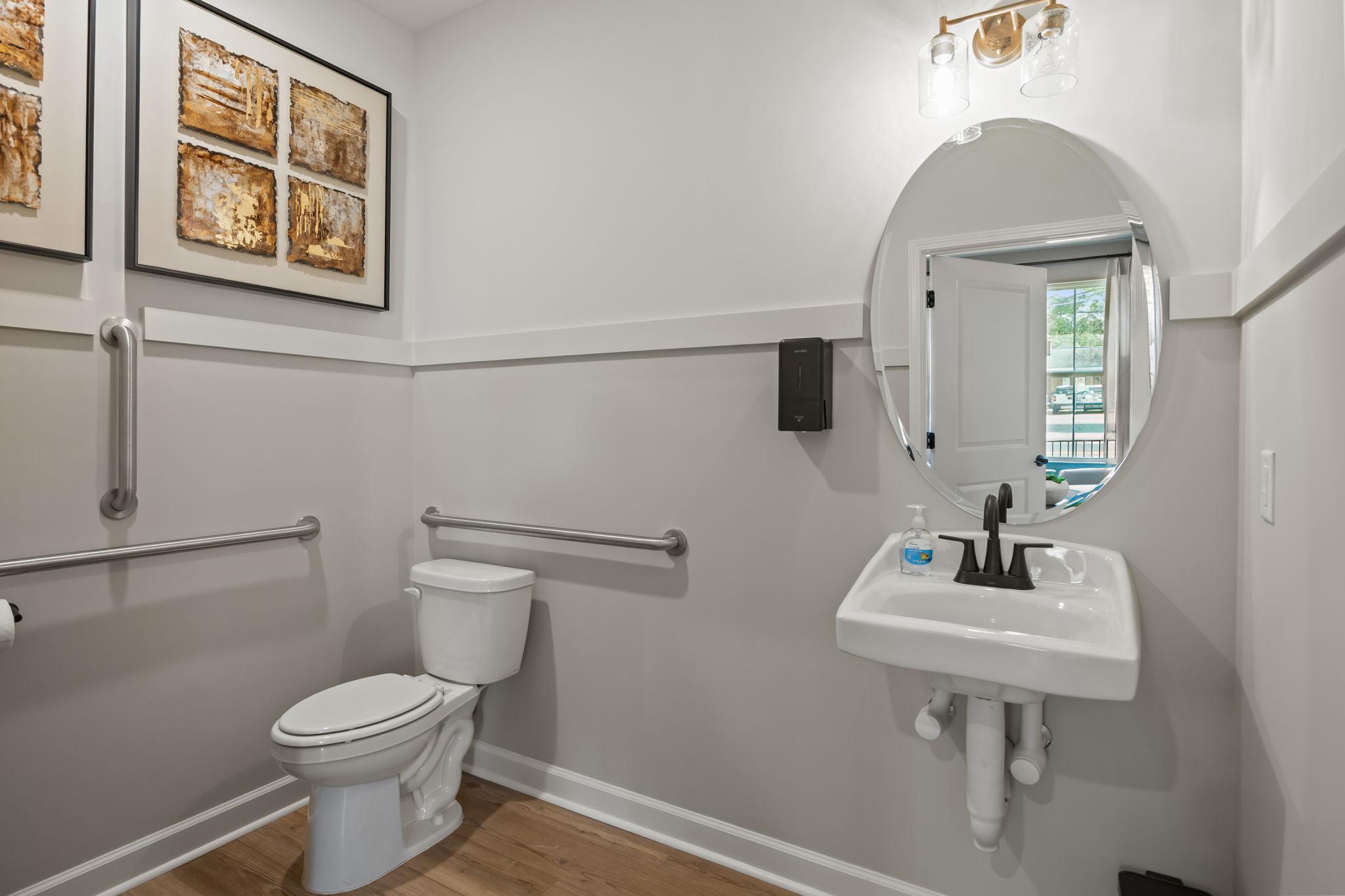 Accessible powder room in The Mitchell interior, featuring a wall-mounted sink, oval mirror, and safety grab bars.