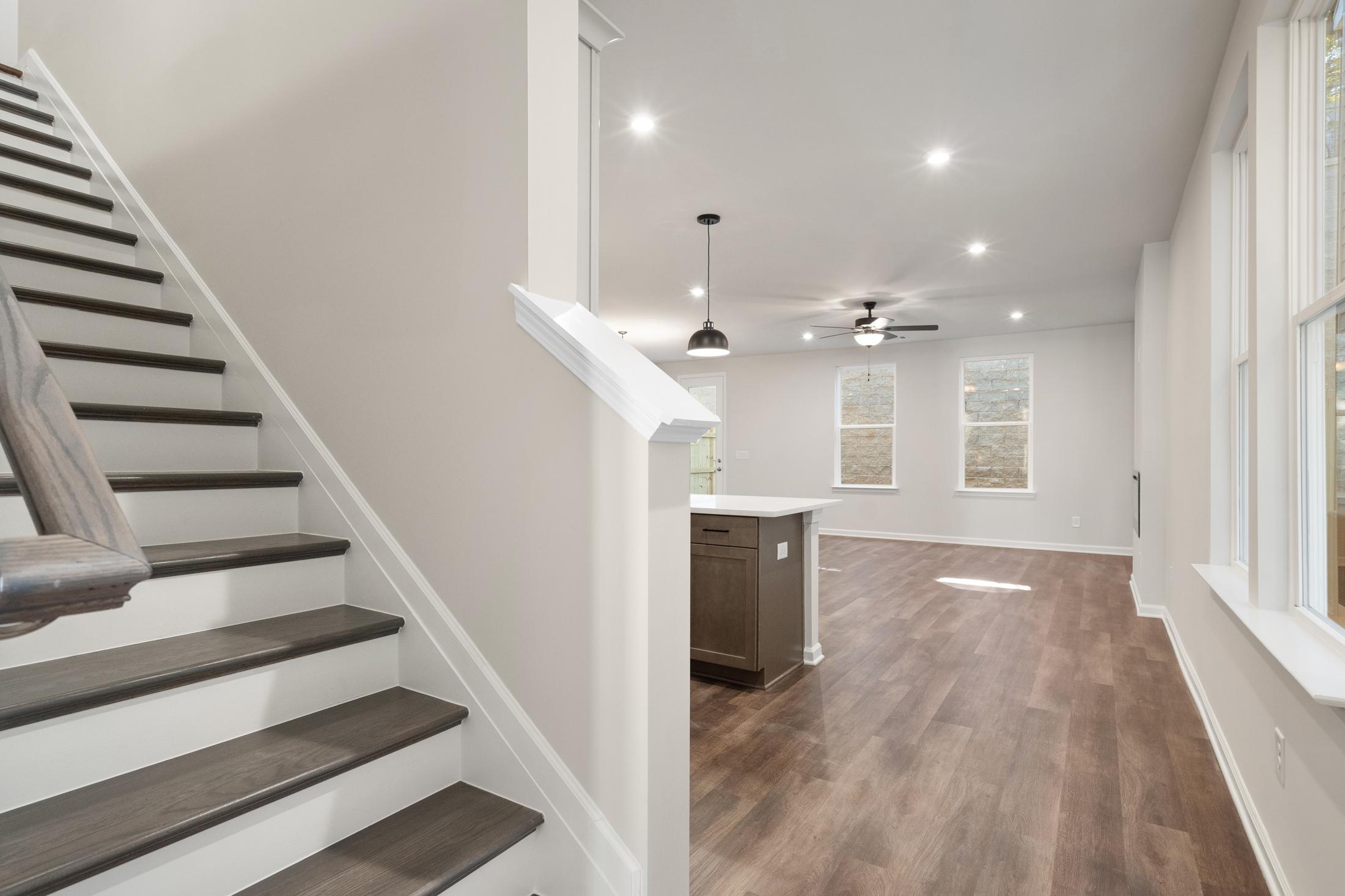 Spacious first-floor open layout of The Cary B with dark wood staircase, white kitchen island, hardwood floors, and large windows