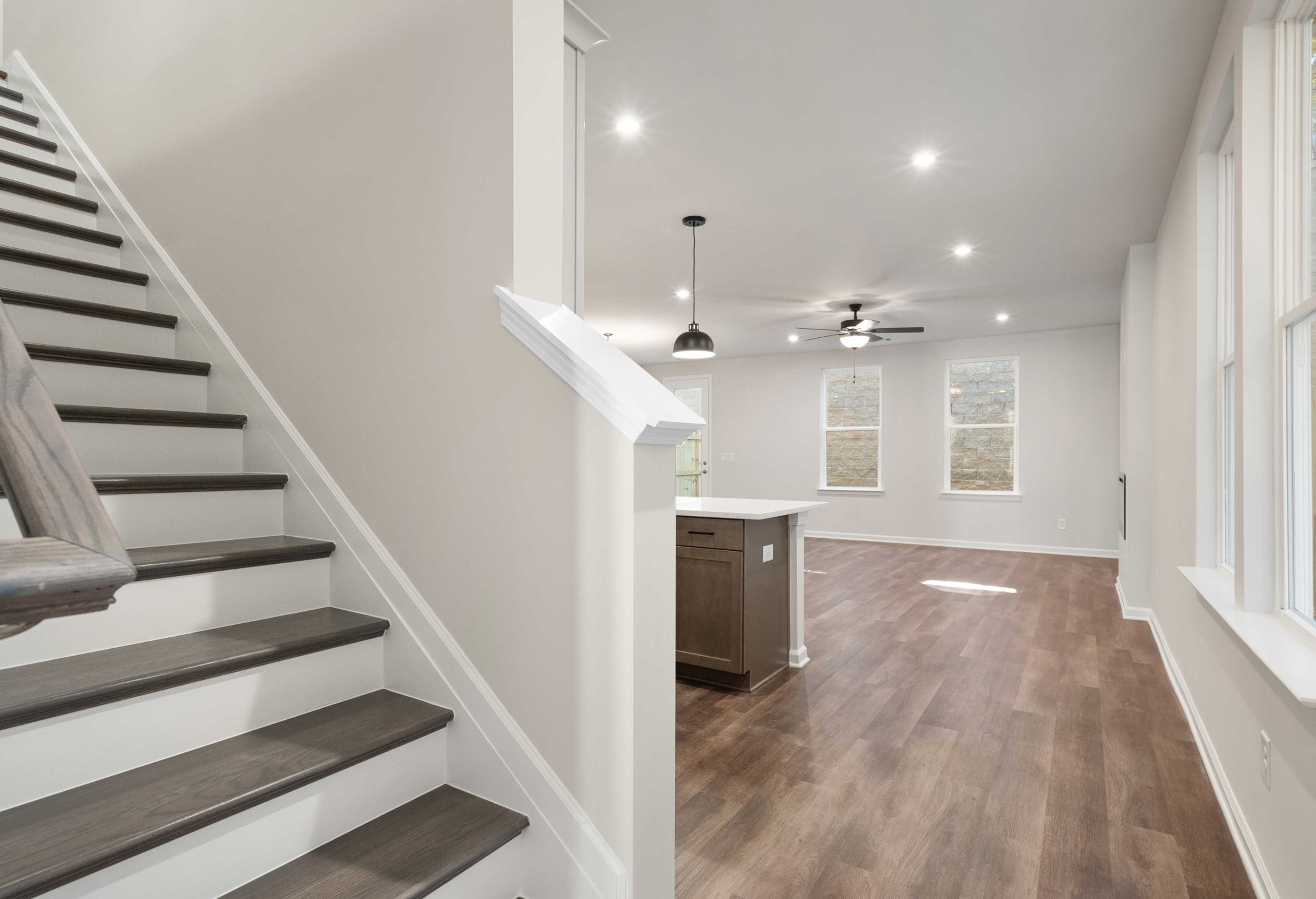 Spacious first-floor open layout of The Cary B with dark wood staircase, white kitchen island, hardwood floors, and large windows