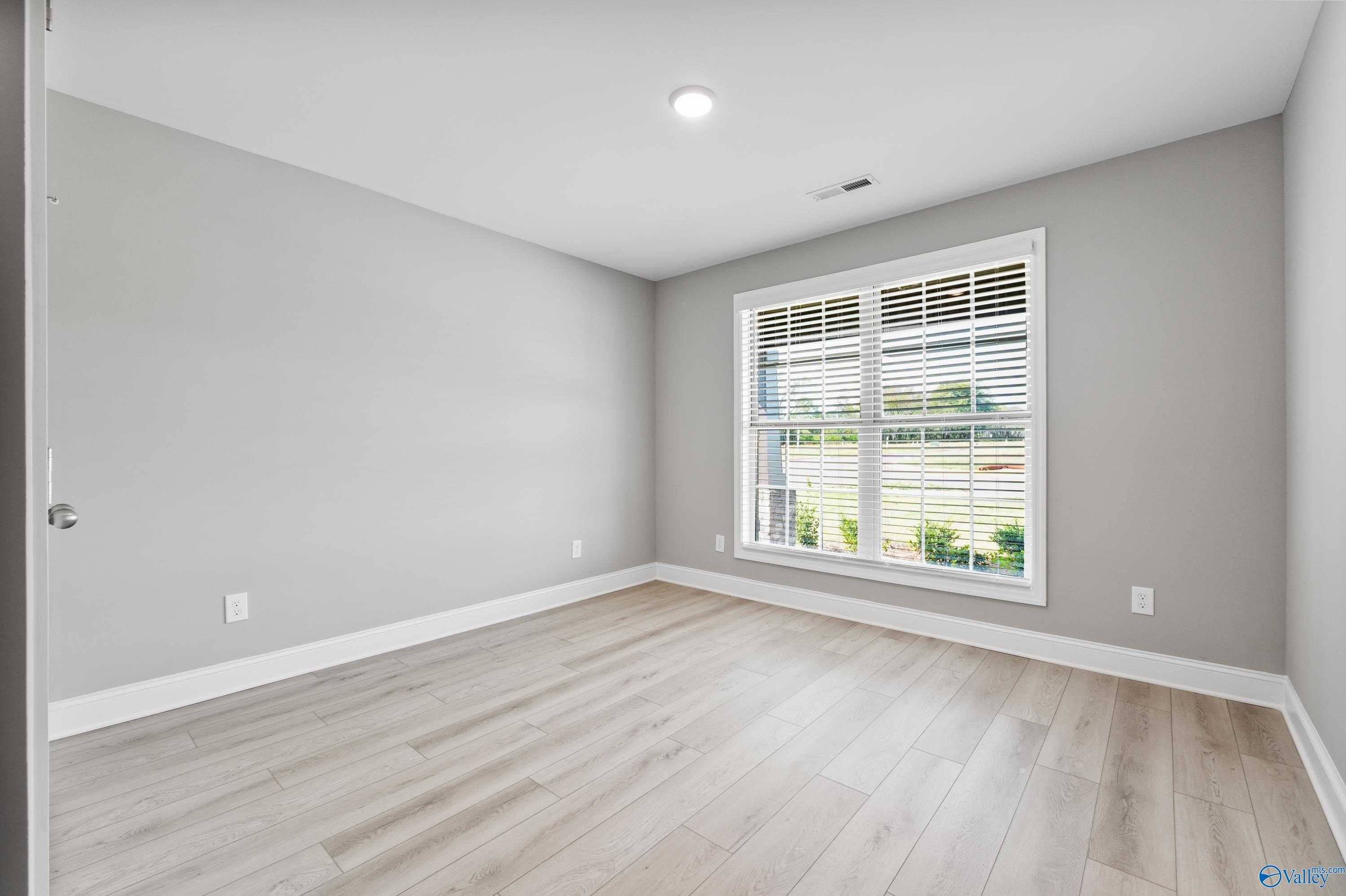 Bright empty bedroom featuring gray walls, light oak floors, and large window with blinds in The Everett B, Athens, AL
