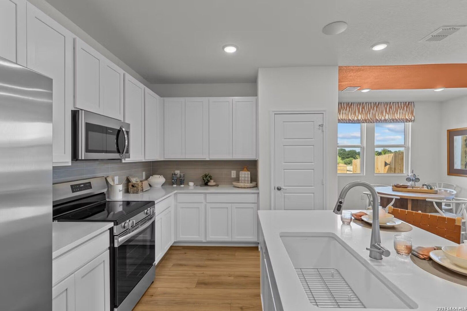 Modern white kitchen with stainless steel appliances, quartz island sink, open to dining area overlooking backyard in Bricewood, San Antonio
