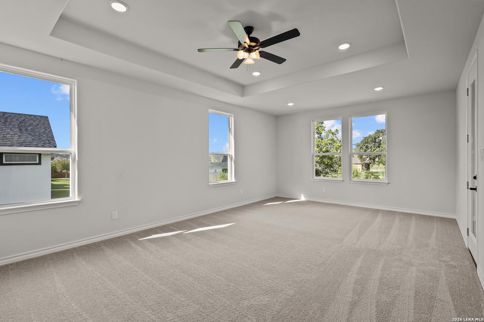 Bright bedroom with tray ceiling, ceiling fan, carpet flooring, and multiple windows in Davidson Homes The Oxford F, Castroville, Texas