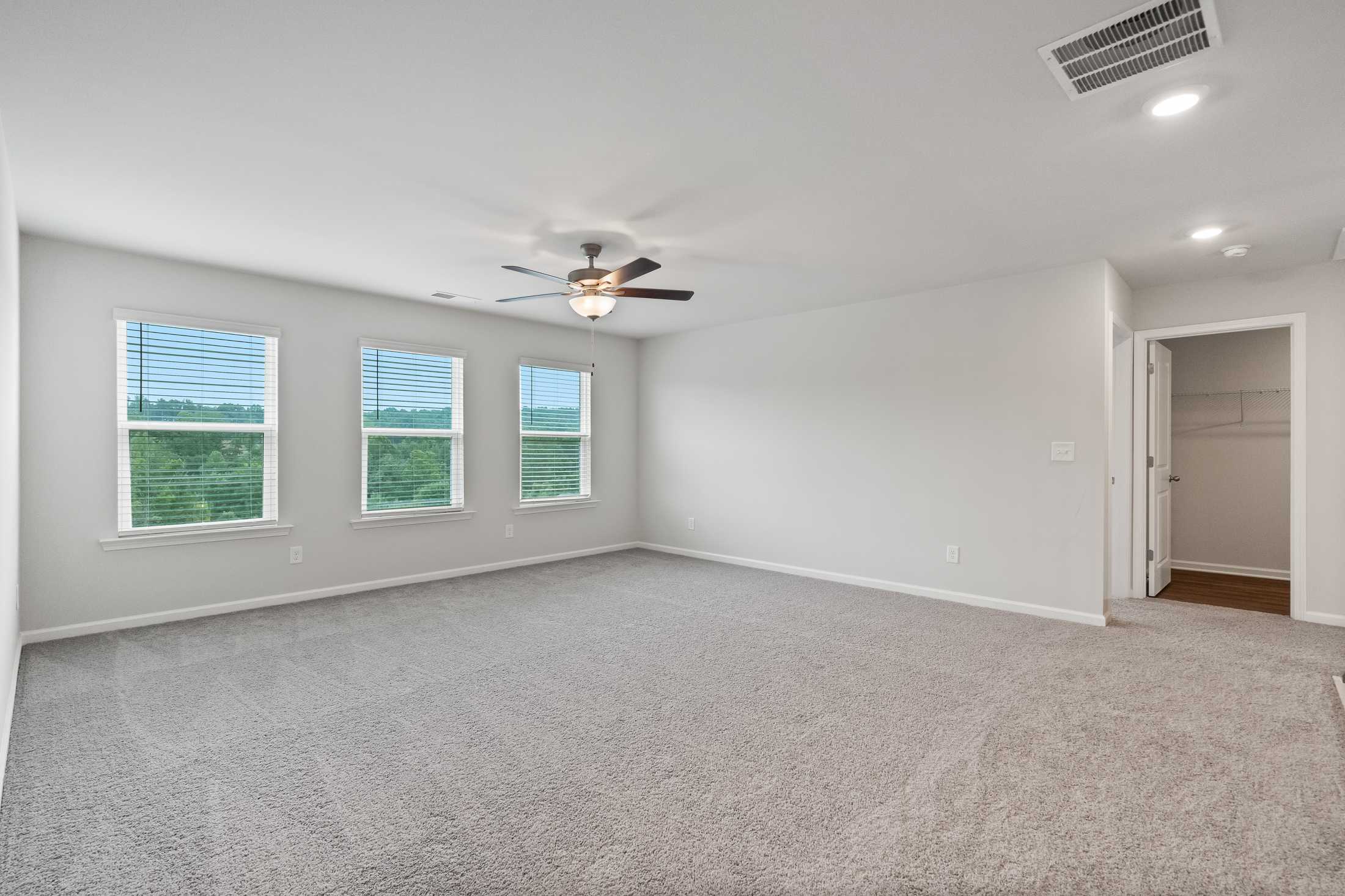 Spacious empty bedroom at Reverie at East Lake in McDonough GA with gray carpet, large windows, blinds, ceiling fan, and walk-in closet