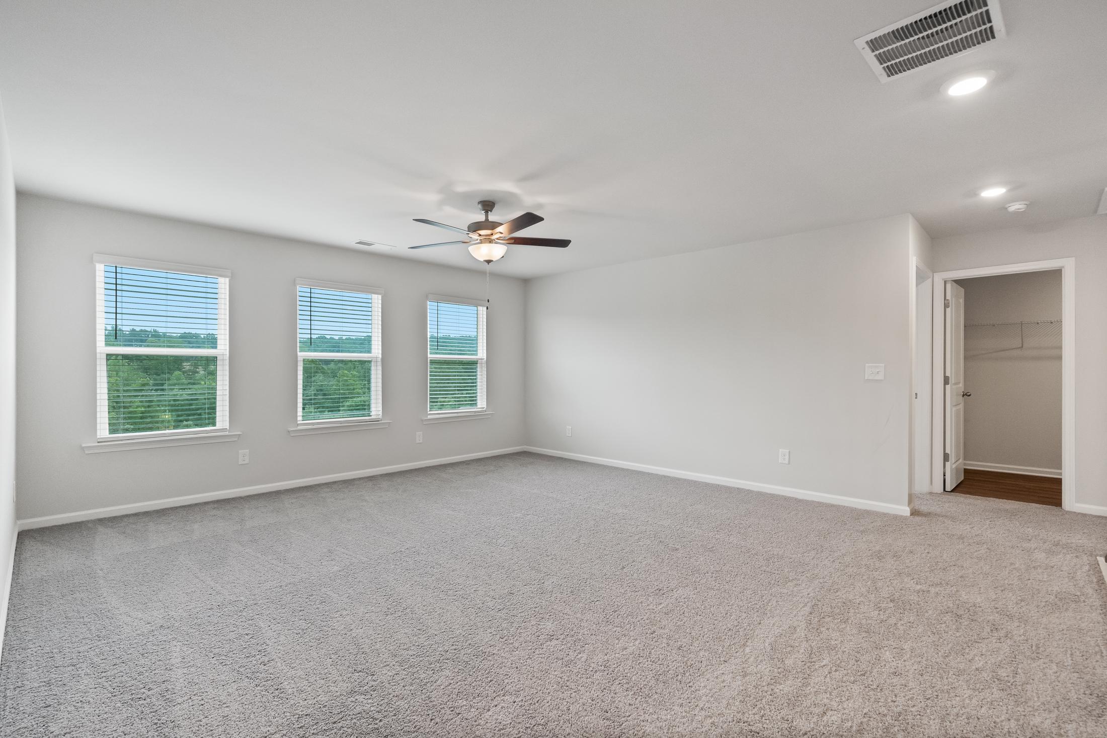 Spacious empty bedroom at Reverie at East Lake in McDonough GA with gray carpet, large windows, blinds, ceiling fan, and walk-in closet
