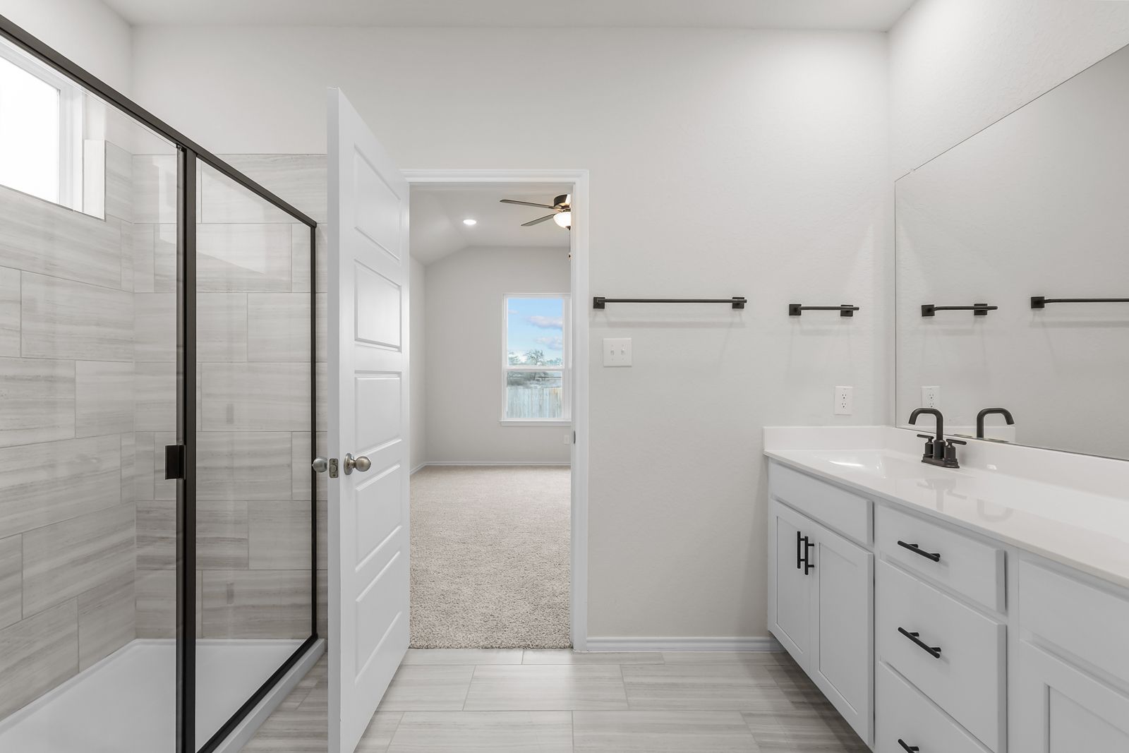 Modern master bathroom in The Sequoia home design featuring a walk-in tiled shower, white vanity, and carpeted bedroom view.