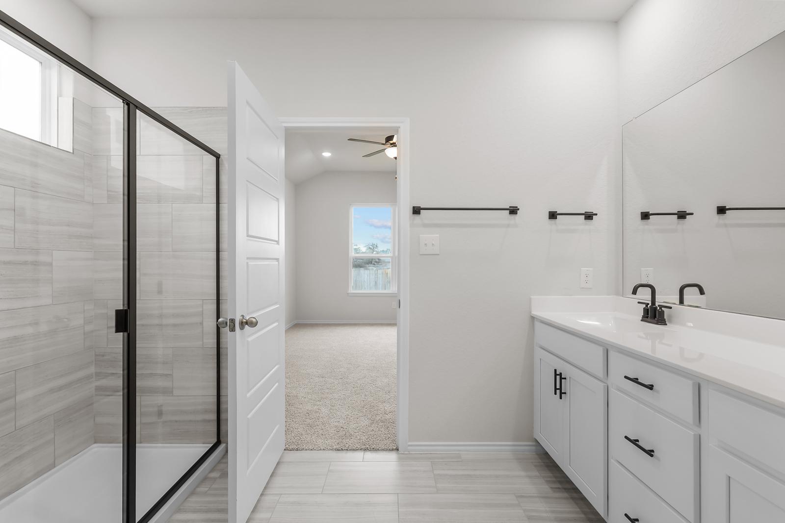 Modern master bathroom in The Sequoia home design featuring a walk-in tiled shower, white vanity, and carpeted bedroom view.