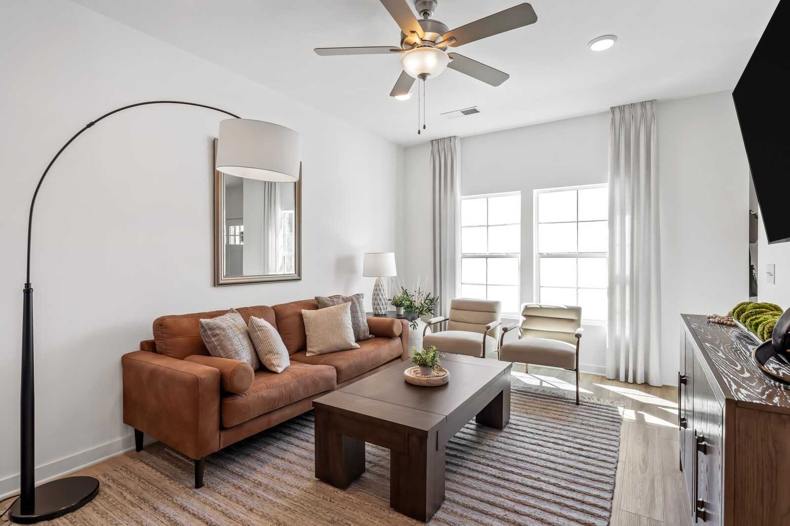Cozy living room with brown leather sofa, wooden coffee table, armchairs, ceiling fan, and large windows in Davidson Homes Cumberland B, Gallatin TN