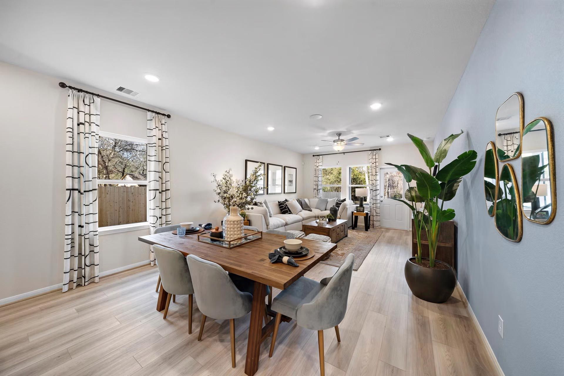 Open-concept dining and living room in modern Cibolo new home with wooden table, upholstered chairs, potted palms, mirrors, and hardwood floors