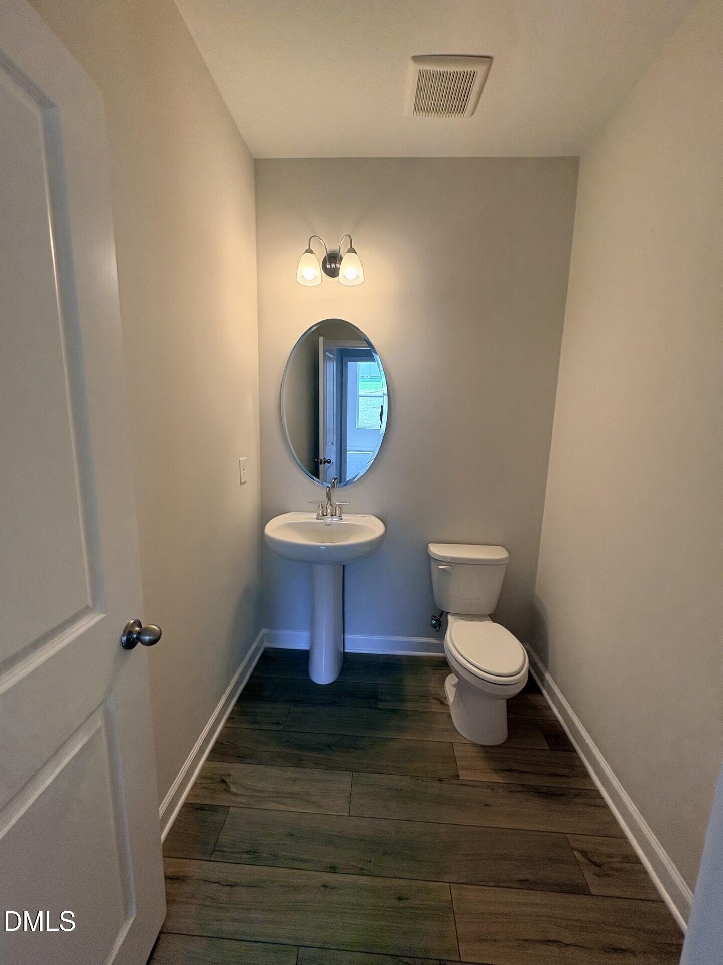 Half-bath with pedestal sink, oval mirror, and wood-look flooring in The Ash C floor plan at Woodland Crossing, Zebulon, NC.