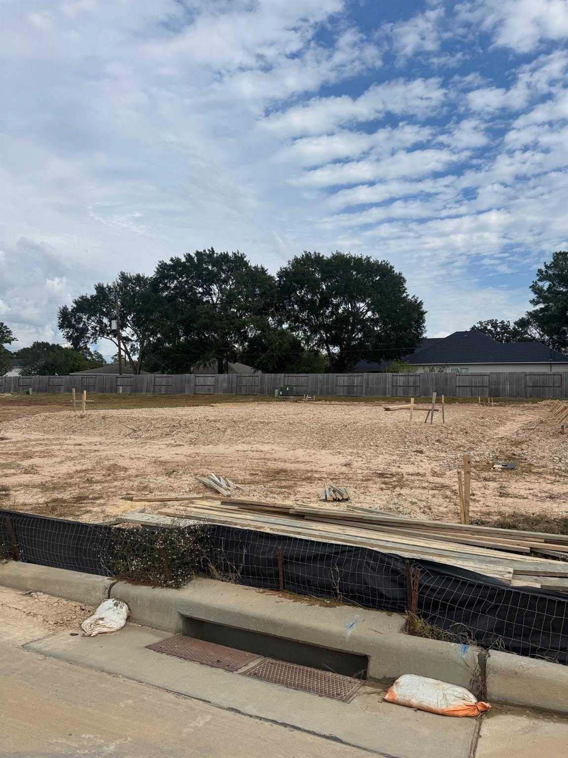 New construction site for Davidson Homes Blanco E in Windmill Estates, Magnolia, Texas: fenced dirt lot, lumber stacks, oak trees backdrop