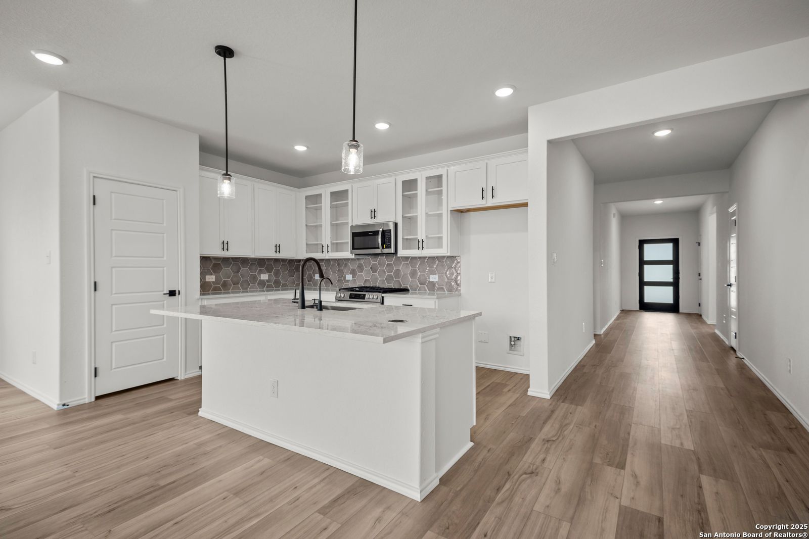 Modern white kitchen with granite island, stainless appliances, and open wood-floor hallway in The Daphne K home, San Antonio