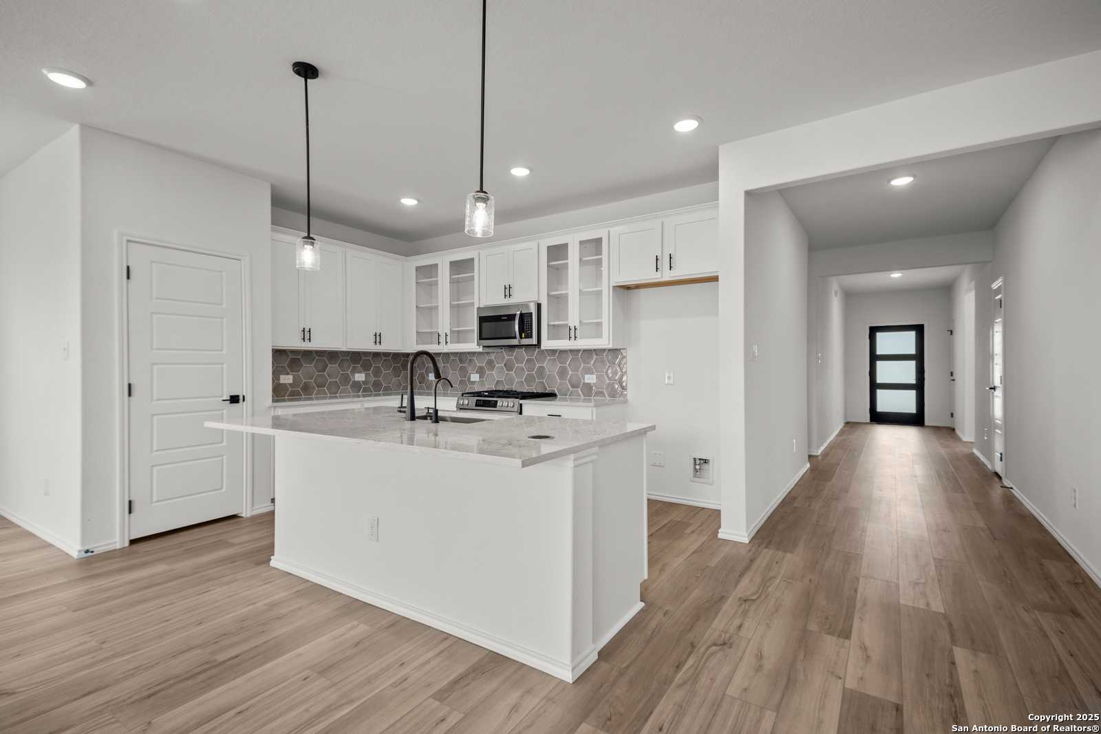 Modern white kitchen with granite island, stainless appliances, and open wood-floor hallway in The Daphne K home, San Antonio