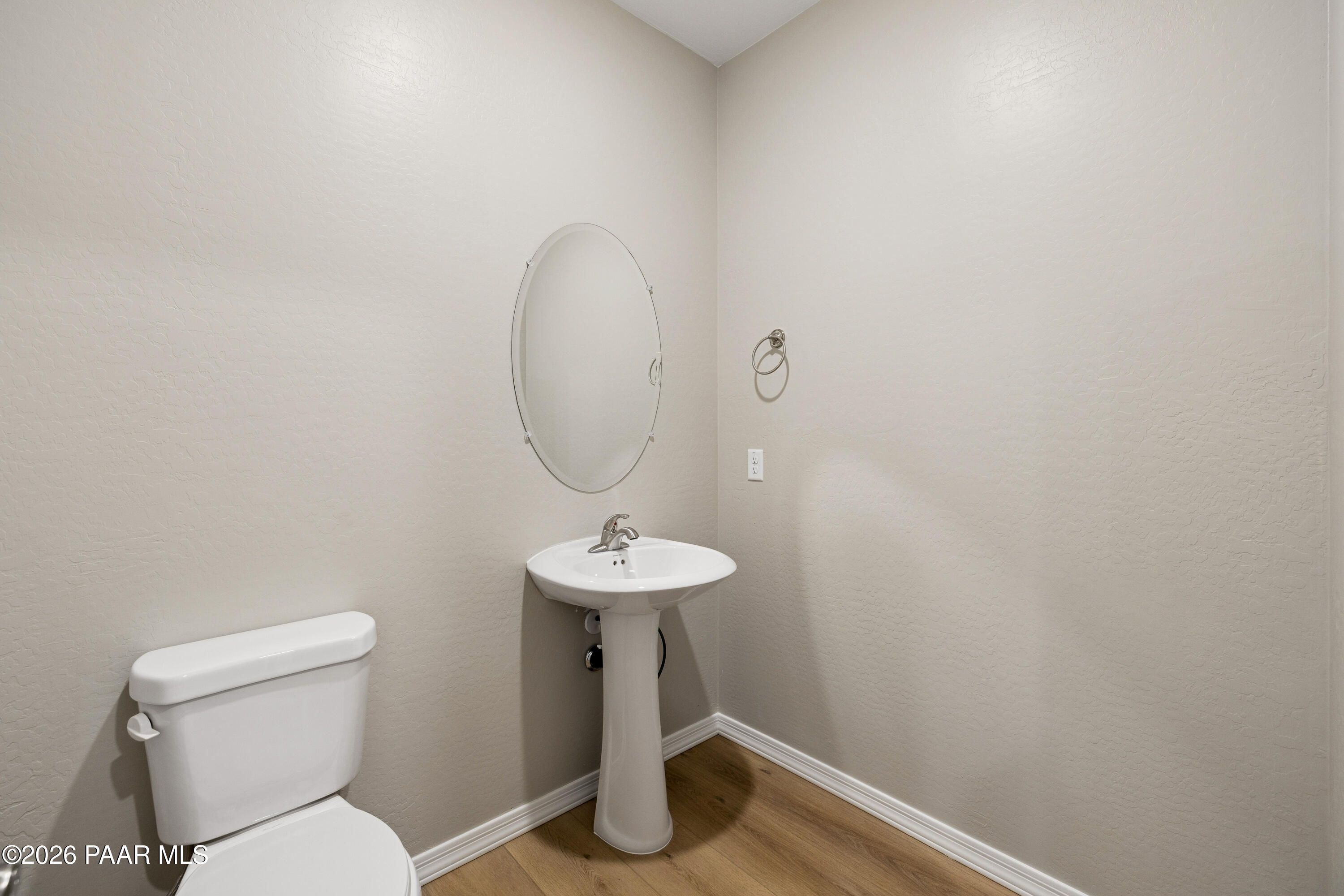 Modern half bathroom featuring pedestal sink, round mirror, white toilet, beige walls, and hardwood floors in Davidson Homes The Harmony A, Prescott Valley, AZ