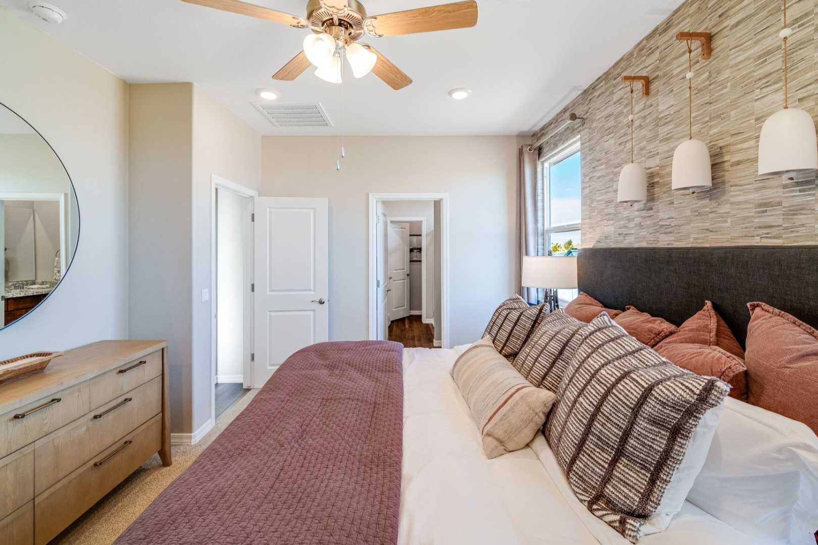 Spacious master bedroom in The Savannah C with king bed, plush pillows, wooden dresser, round mirror, and textured wall accents