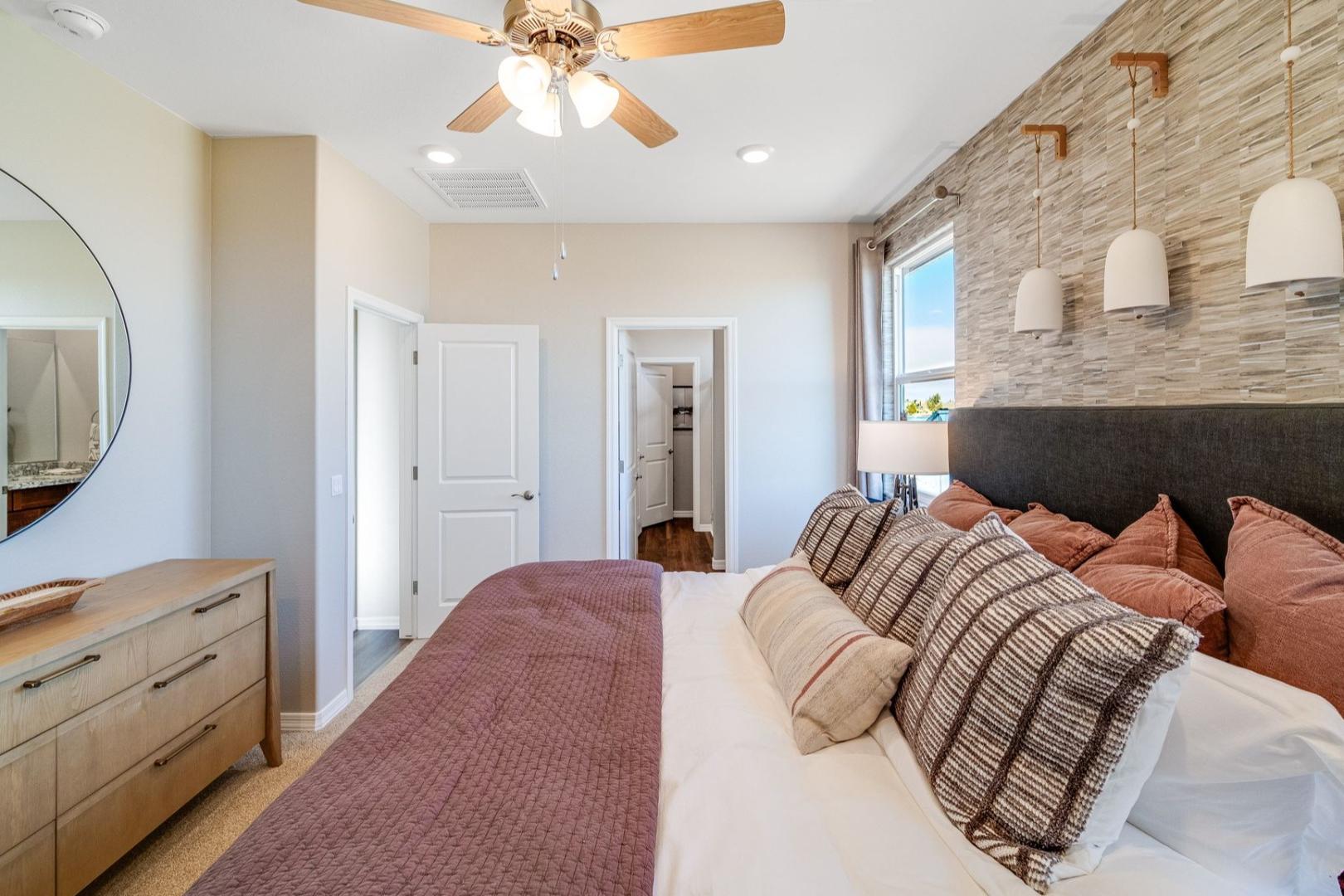 Spacious master bedroom in The Savannah B featuring king bed with plush pillows, wooden dresser, round mirror, and pendant lights