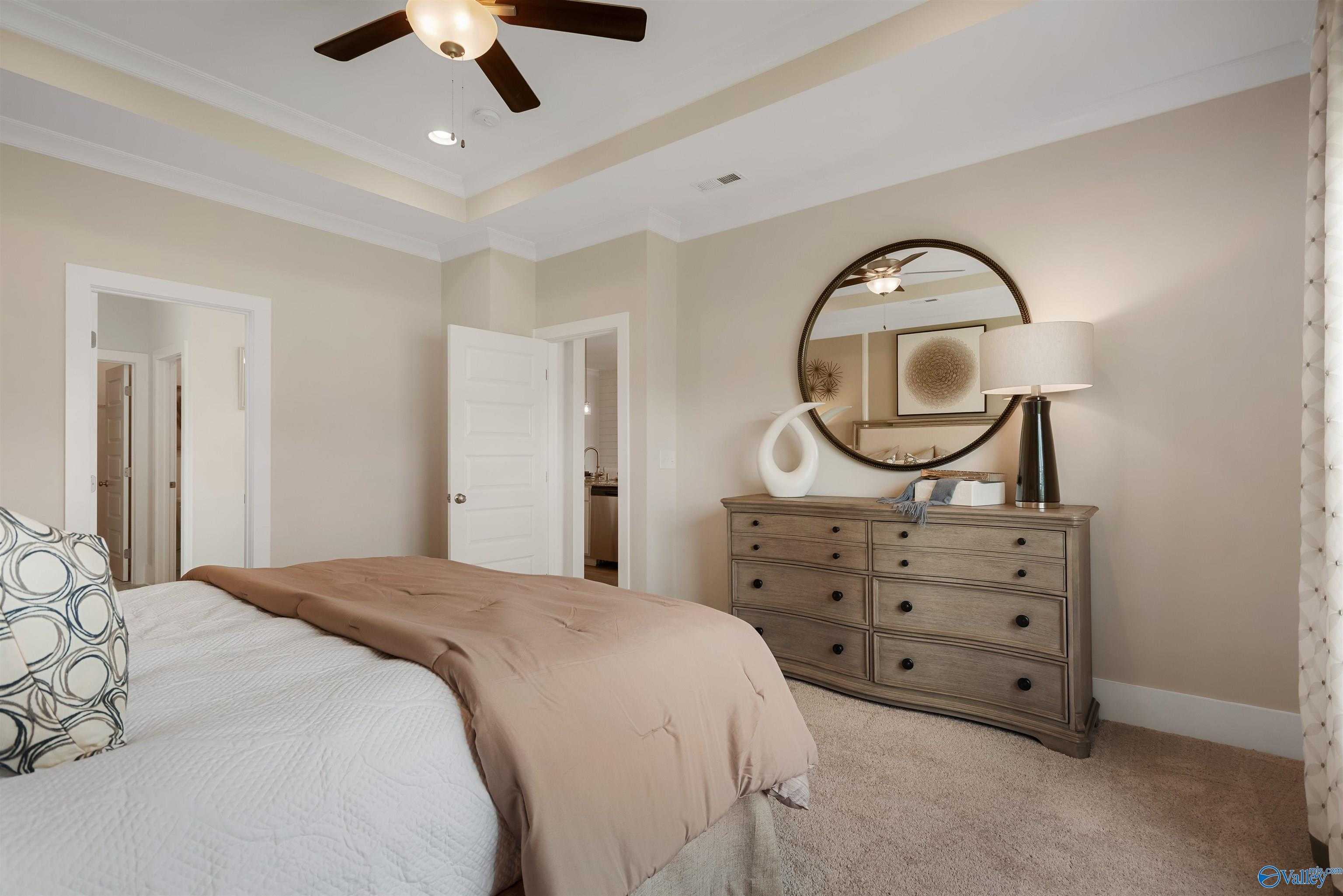 Serene master bedroom with tray ceiling, ceiling fan, round mirror over wooden dresser, neutral bed in Davidson Homes The Everett, New Market AL