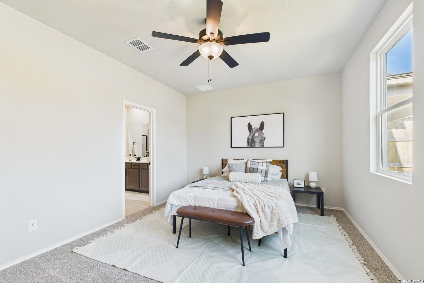 Cozy bedroom with neutral bedding, ceiling fan, horse artwork, and adjacent bathroom in Davidson Homes Douglas E, Bricewood, San Antonio