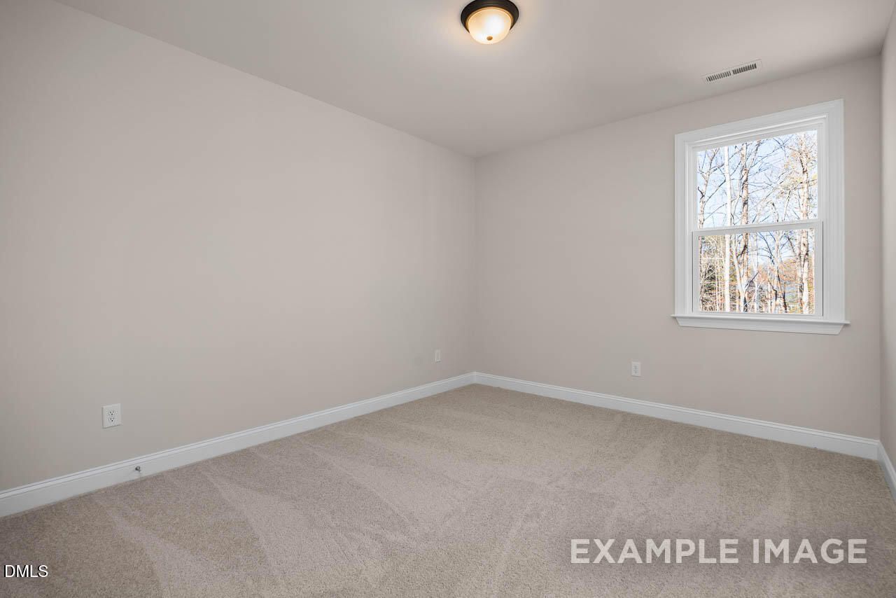Spacious empty bedroom with neutral beige walls, plush carpet, and window overlooking trees in The Crawford D, Angier, NC
