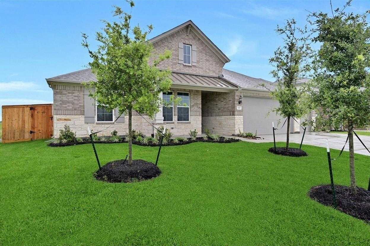 Beige brick single-story home with 3-car garage, lush green lawn, young trees, and mulch beds in Lago Mar, Texas City