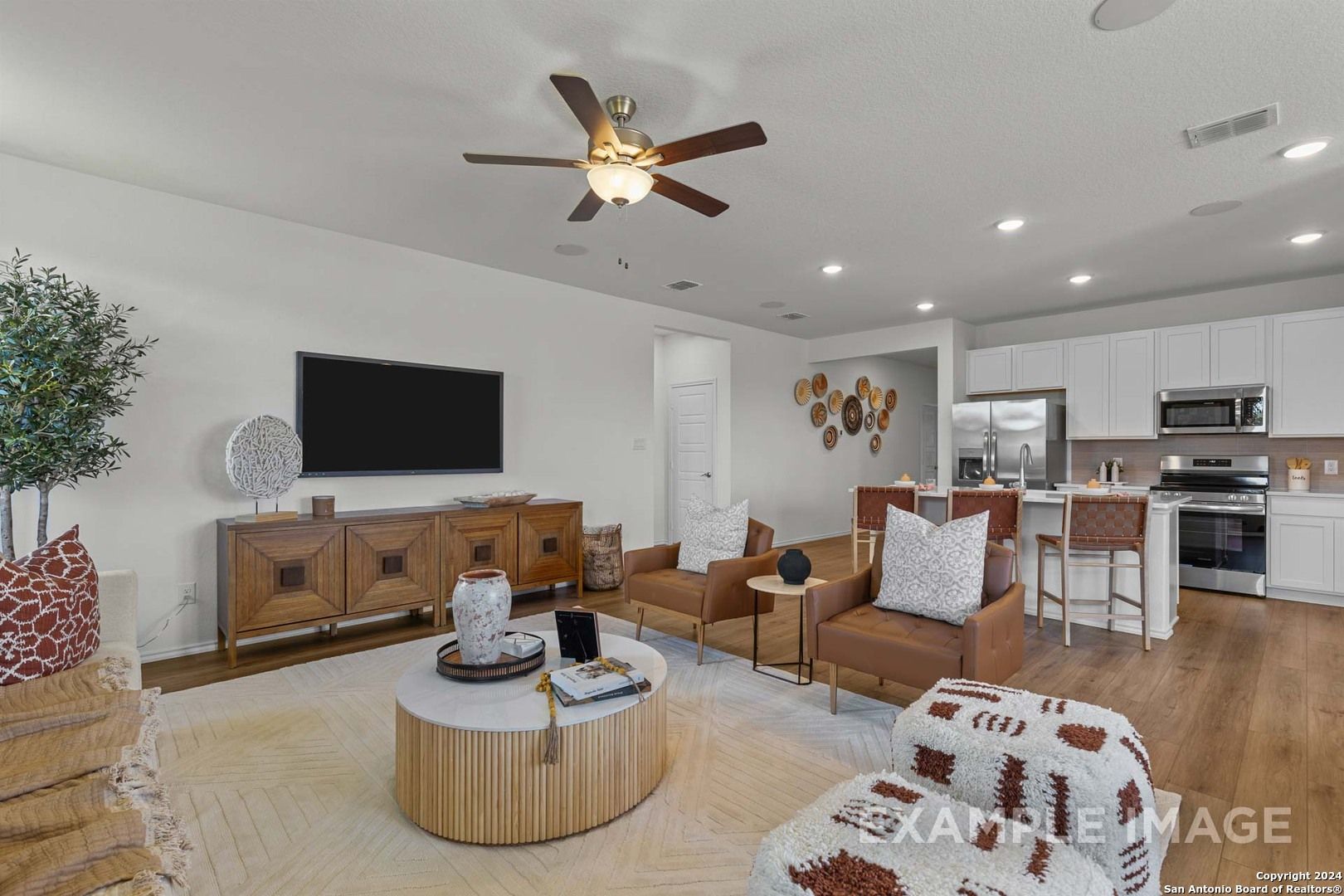 Open-concept living room with white kitchen, beige sofa, wall TV, ceiling fan in The Daphne K by Davidson Homes, San Antonio