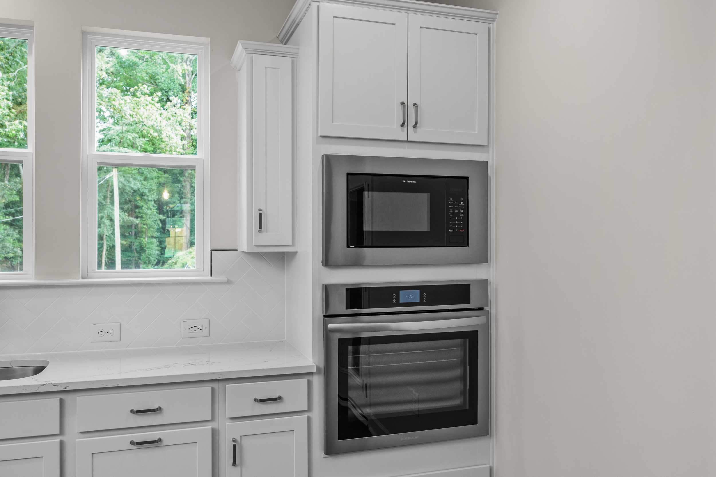 Modern kitchen in The Aspen by Davidson Homes with white shaker cabinets, stainless steel built-in microwave and oven, white quartz counters, large windows to greenery