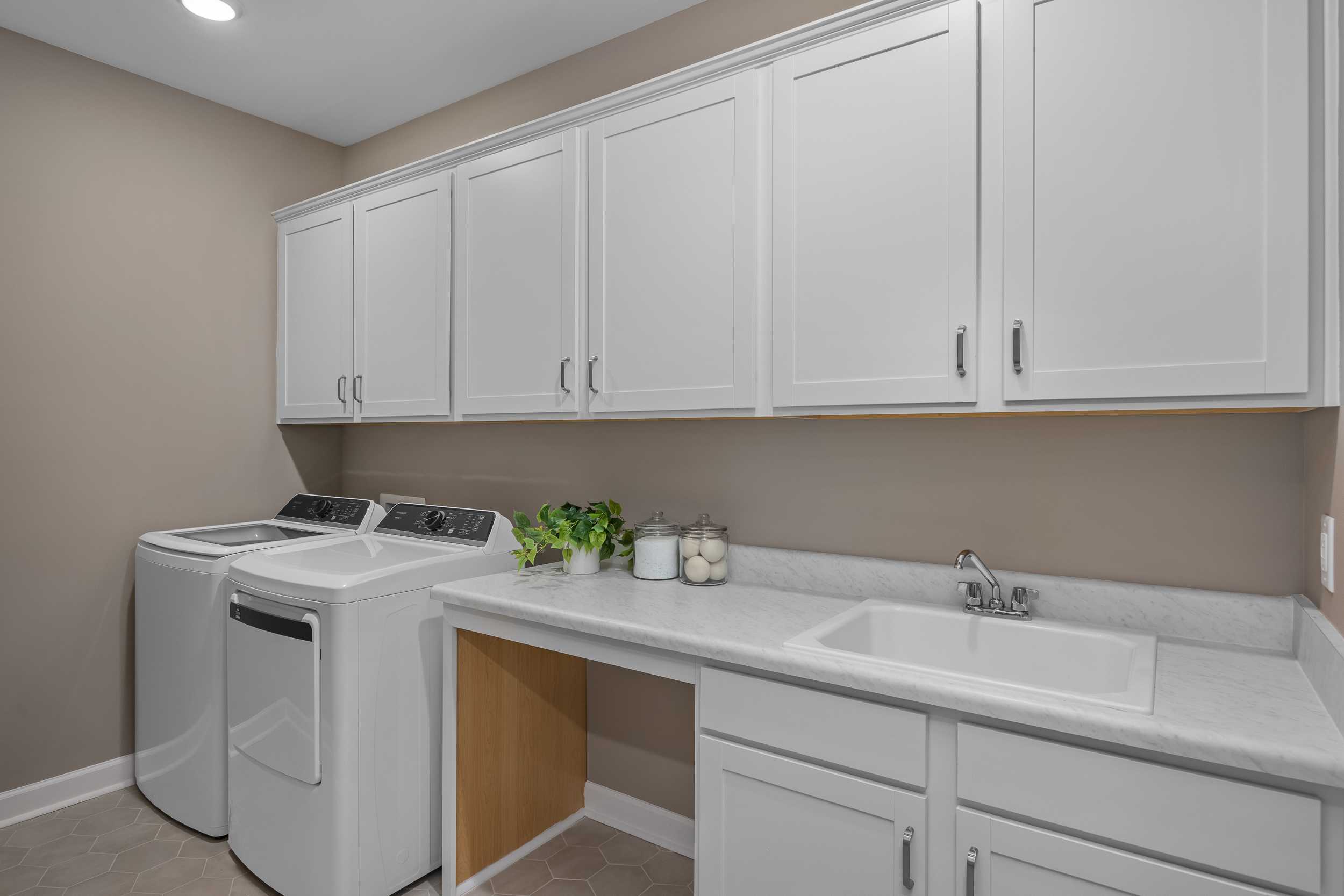 Spacious laundry room in The Hickory II E featuring white washer dryer, shaker cabinets, quartz countertop, utility sink, and beige walls