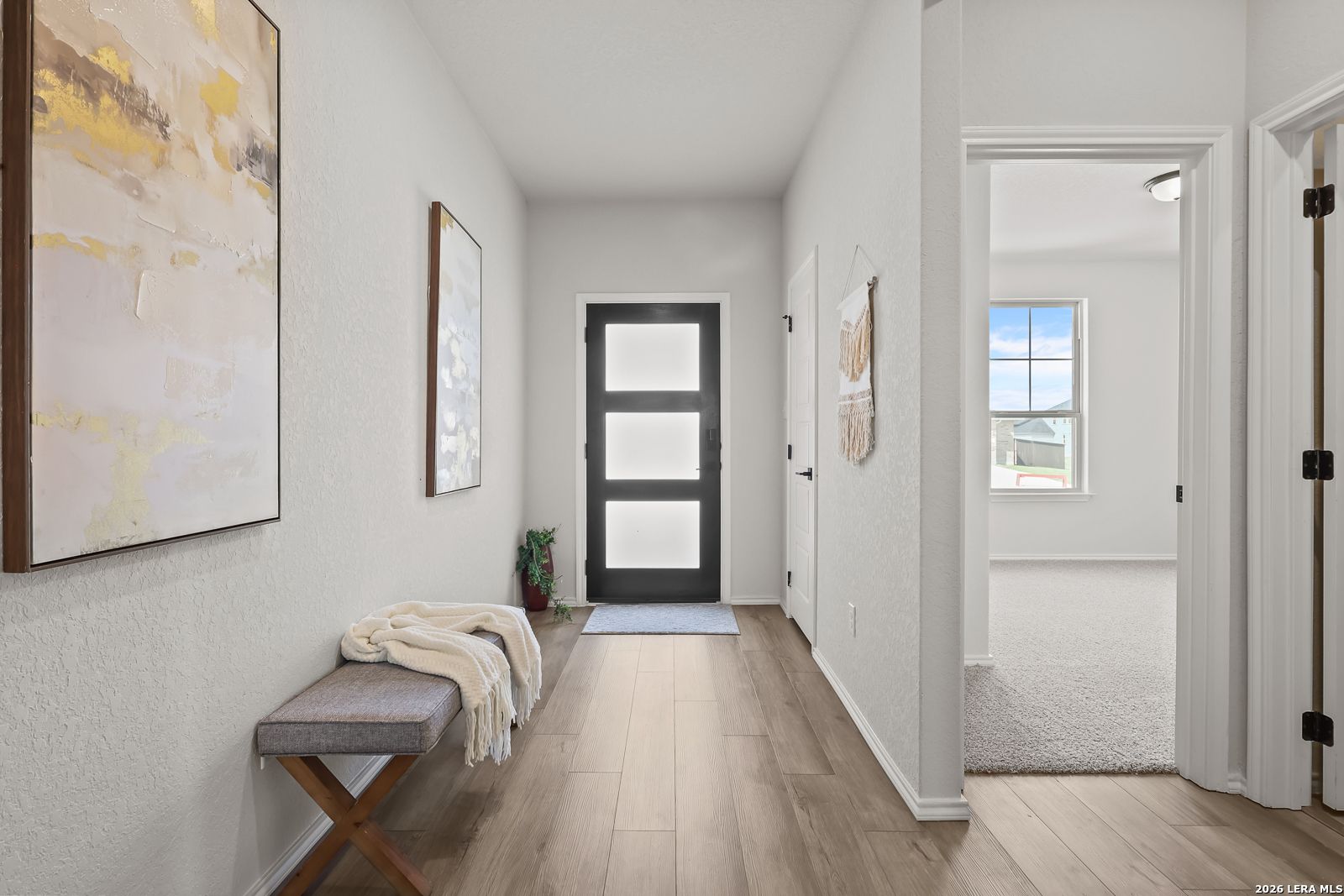 Bright entryway hallway with light wood floors, abstract wall art, bench, and glass front door in Davidson Homes The Daphne H, Seguin, Texas