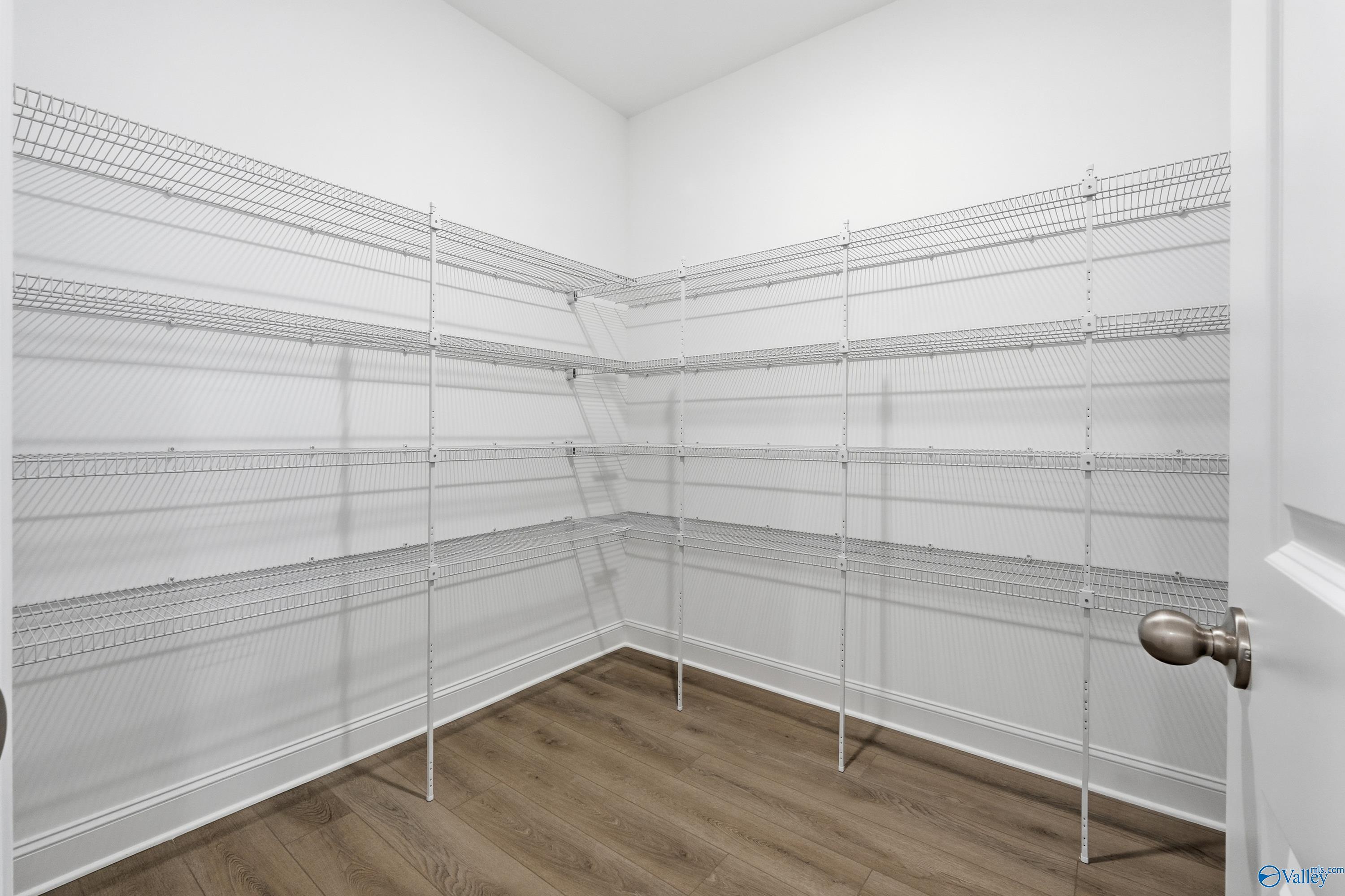 Spacious corner walk-in pantry with white wire shelving, open door, and wood flooring in The Finleigh by Davidson Homes, Harvest, Alabama