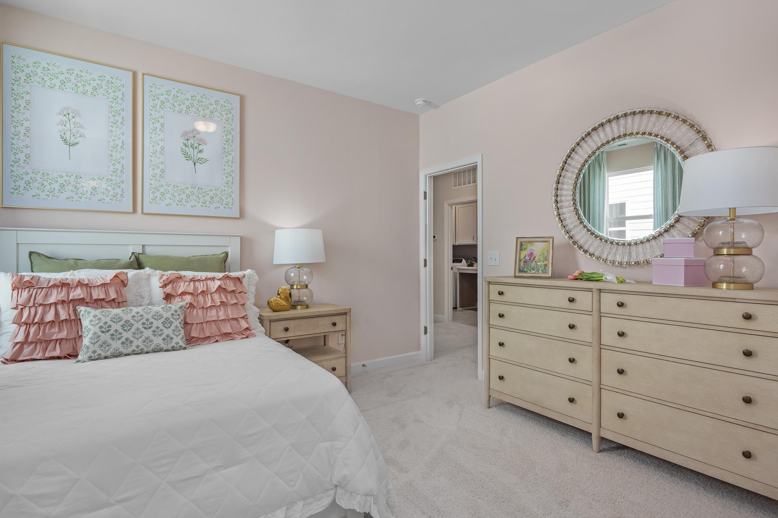 Cozy master bedroom in The Hickory II with blush pink walls, king bed, colorful pillows, wooden dresser, round mirror, and botanical prints