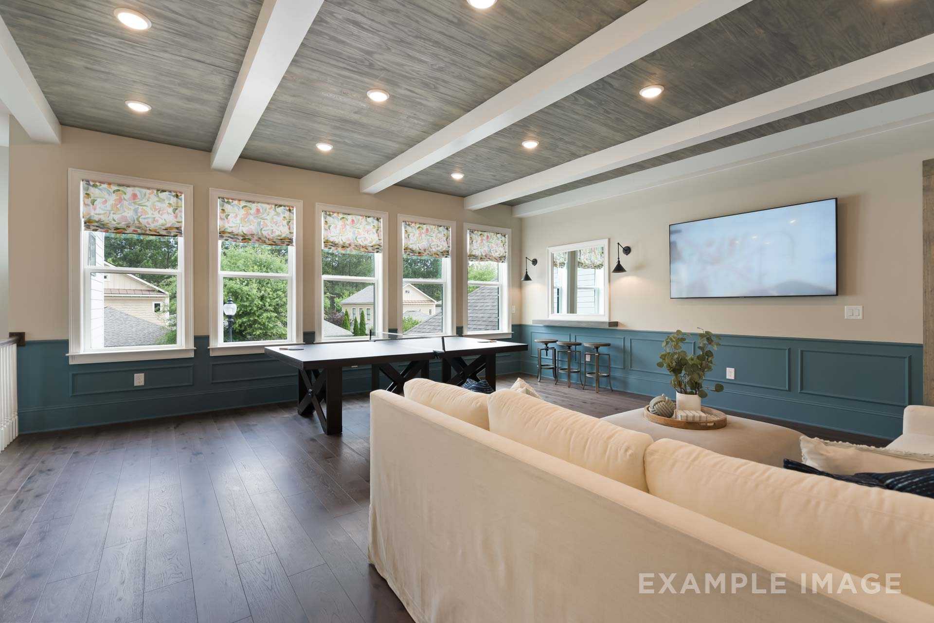 Spacious loft bonus room in The Seaside A with exposed beam ceiling, navy wainscoting, white sofa, game table and large windows