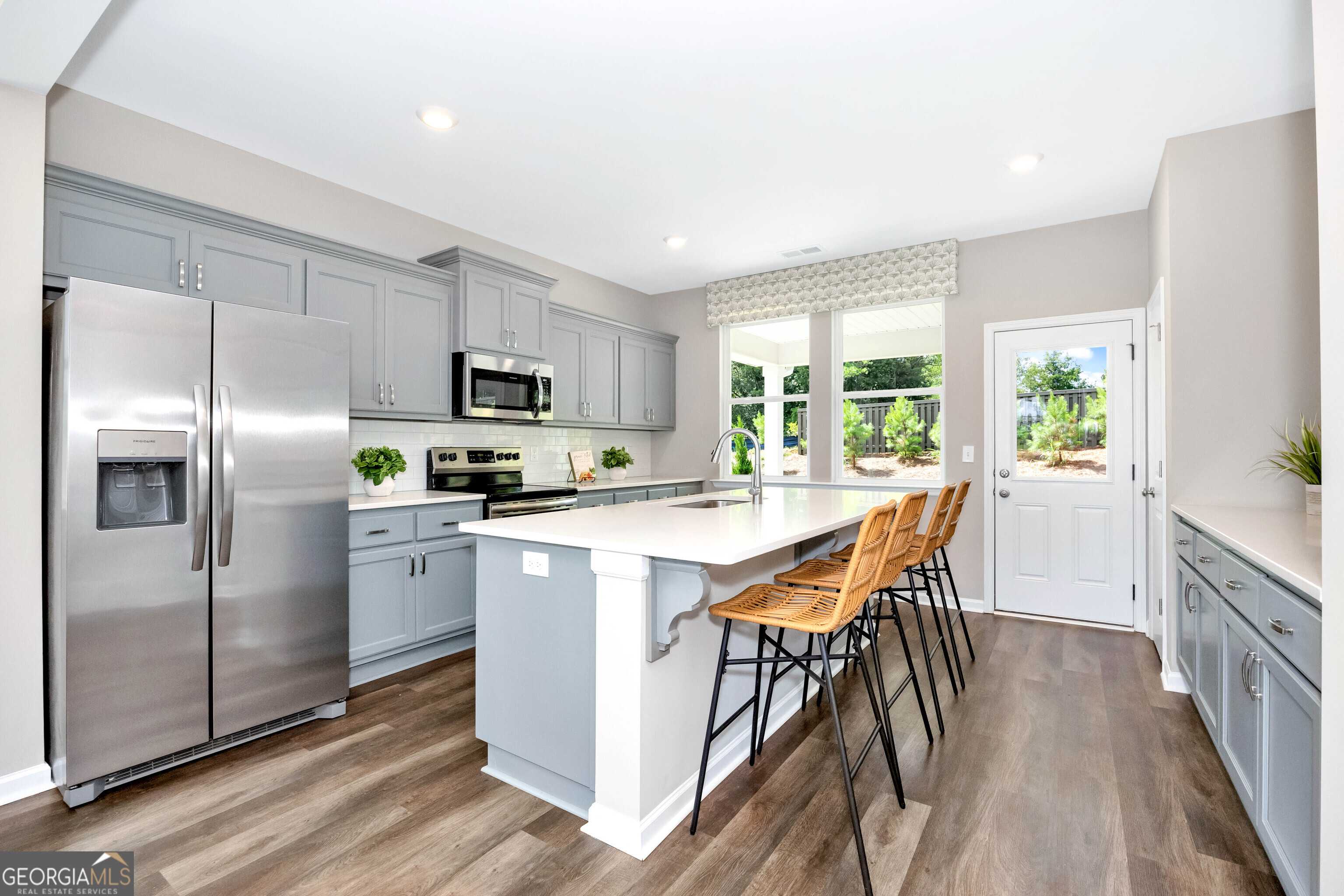 Modern kitchen featuring gray cabinets, stainless steel fridge, microwave, and white island with bar stools in Evermore Homes The Stella, Perry, Georgia