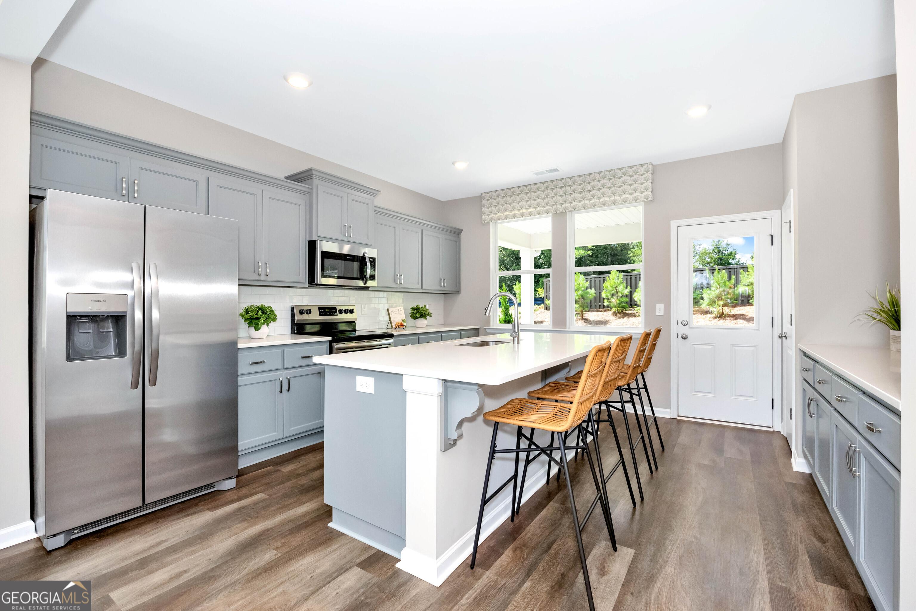 Modern kitchen featuring gray cabinets, stainless steel fridge, microwave, and white island with bar stools in Evermore Homes The Stella, Perry, Georgia