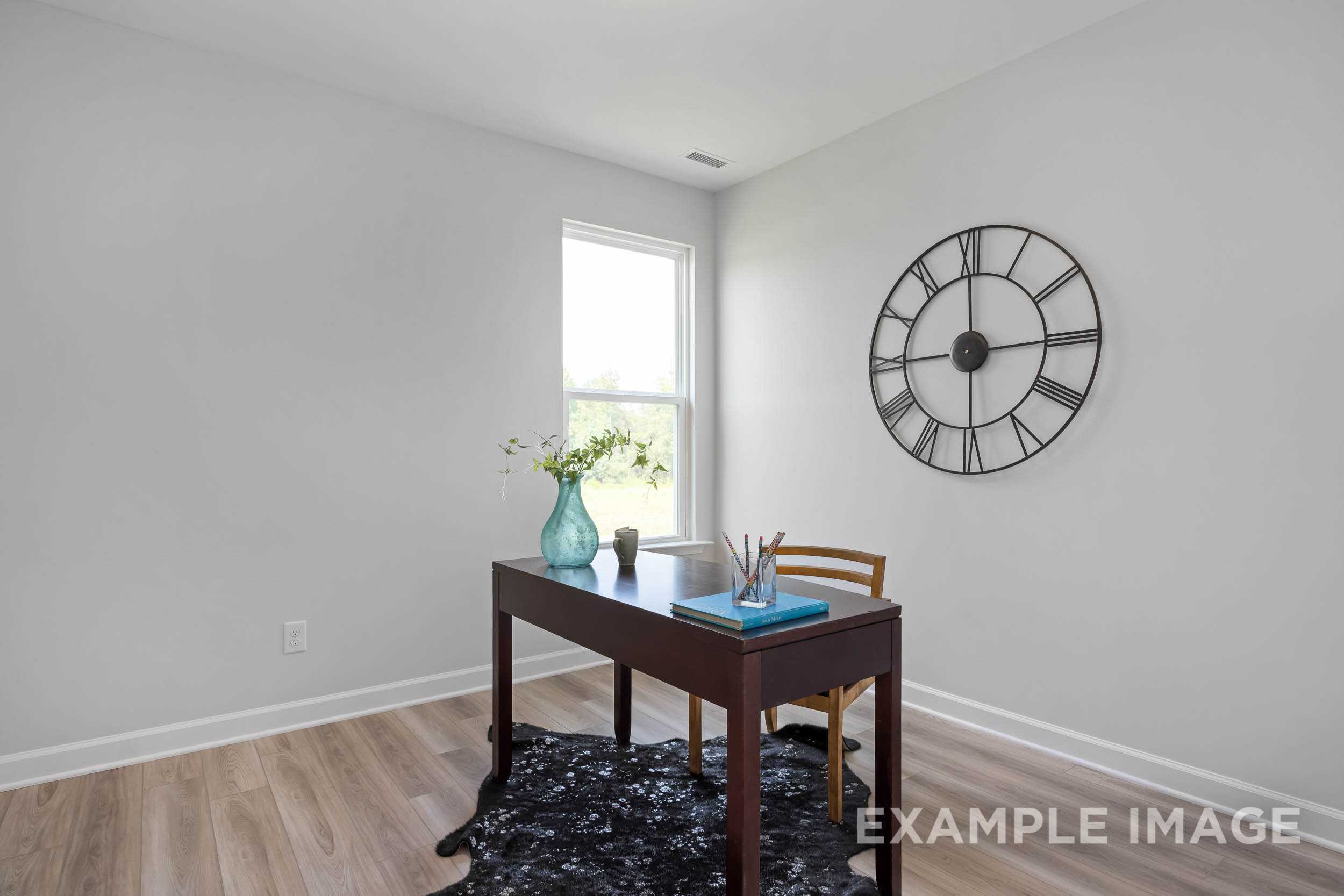 Cozy home office in The Daphne C featuring wooden desk, blue vase of flowers, large wall clock, and window light