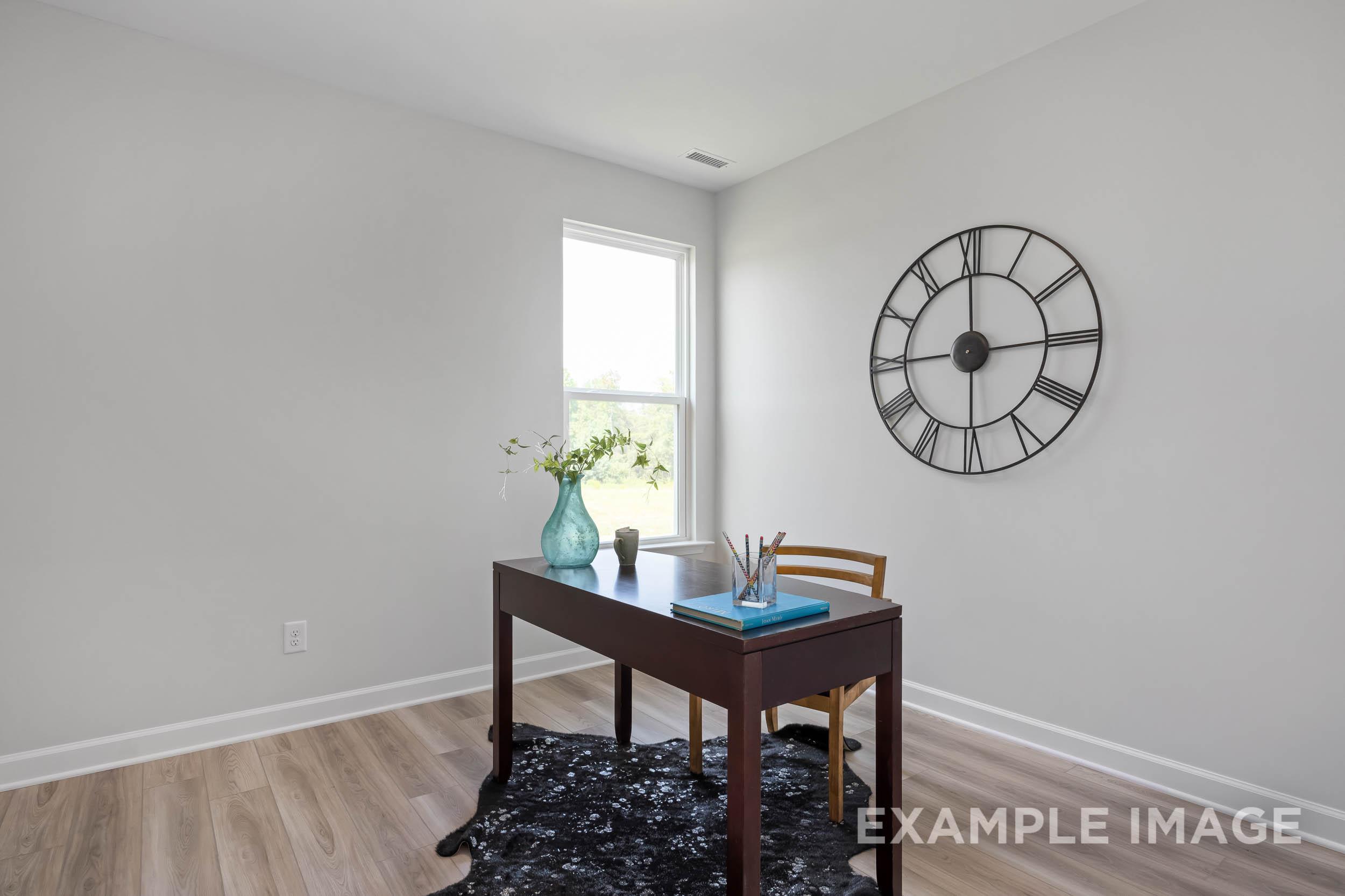Cozy home office in The Daphne C featuring wooden desk, blue vase of flowers, large wall clock, and window light