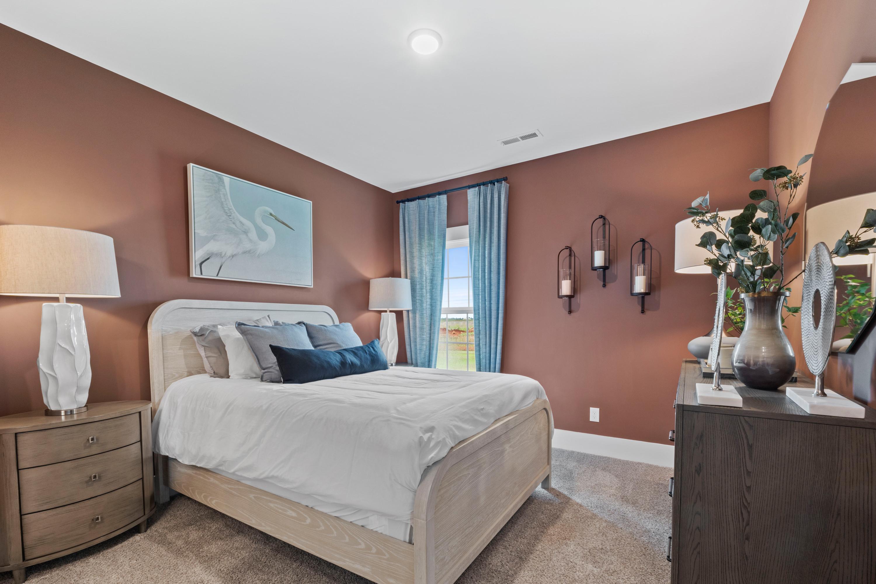 Primary bedroom interior at Creekside in Harvest Alabama featuring terracotta walls, white king bed, egret painting, and wooden accents