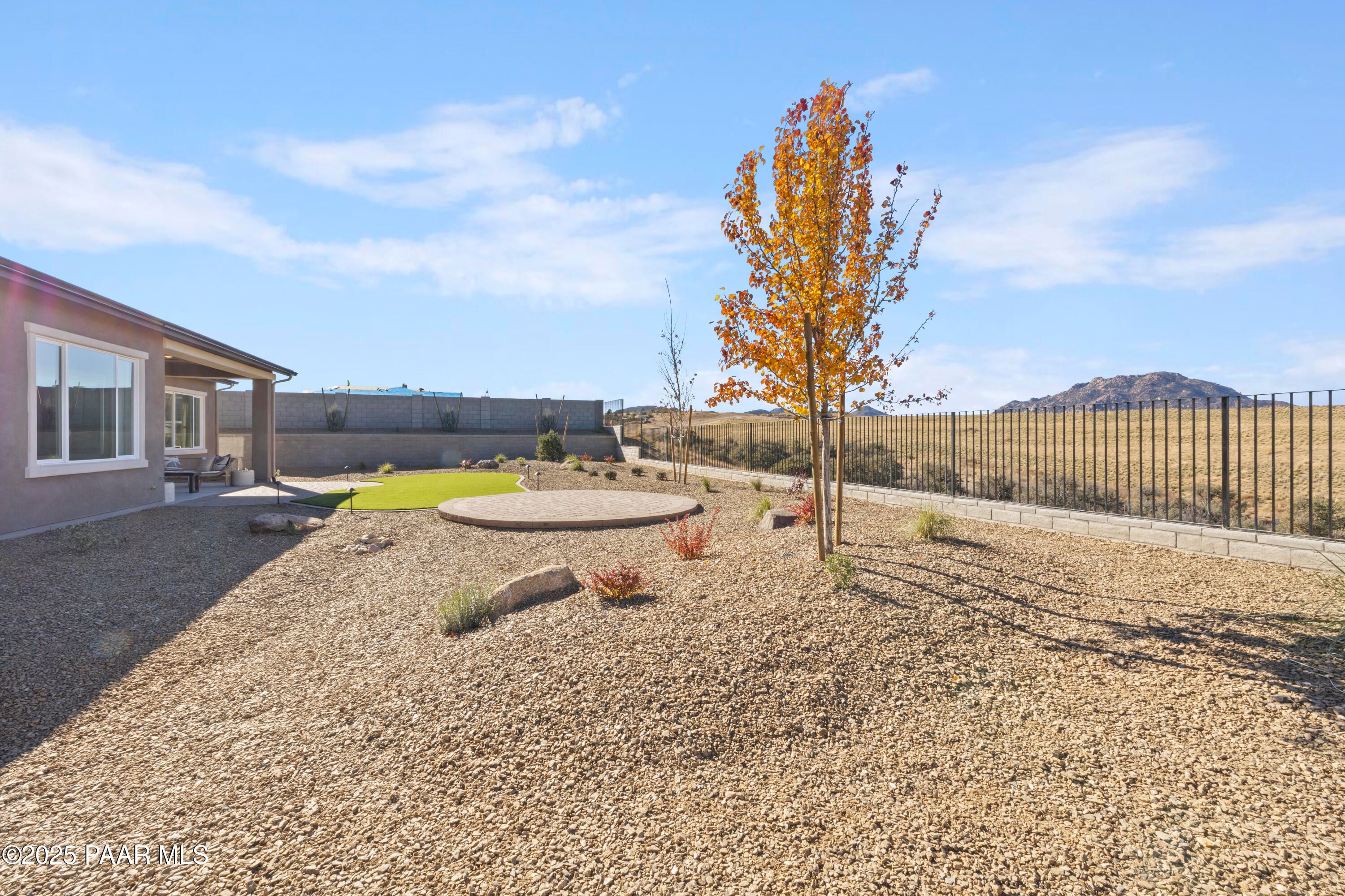 Desert backyard with gravel landscaping, orange autumn tree, patio seating, and mountain views beside single-story home in Hidden Hills, Prescott, Arizona