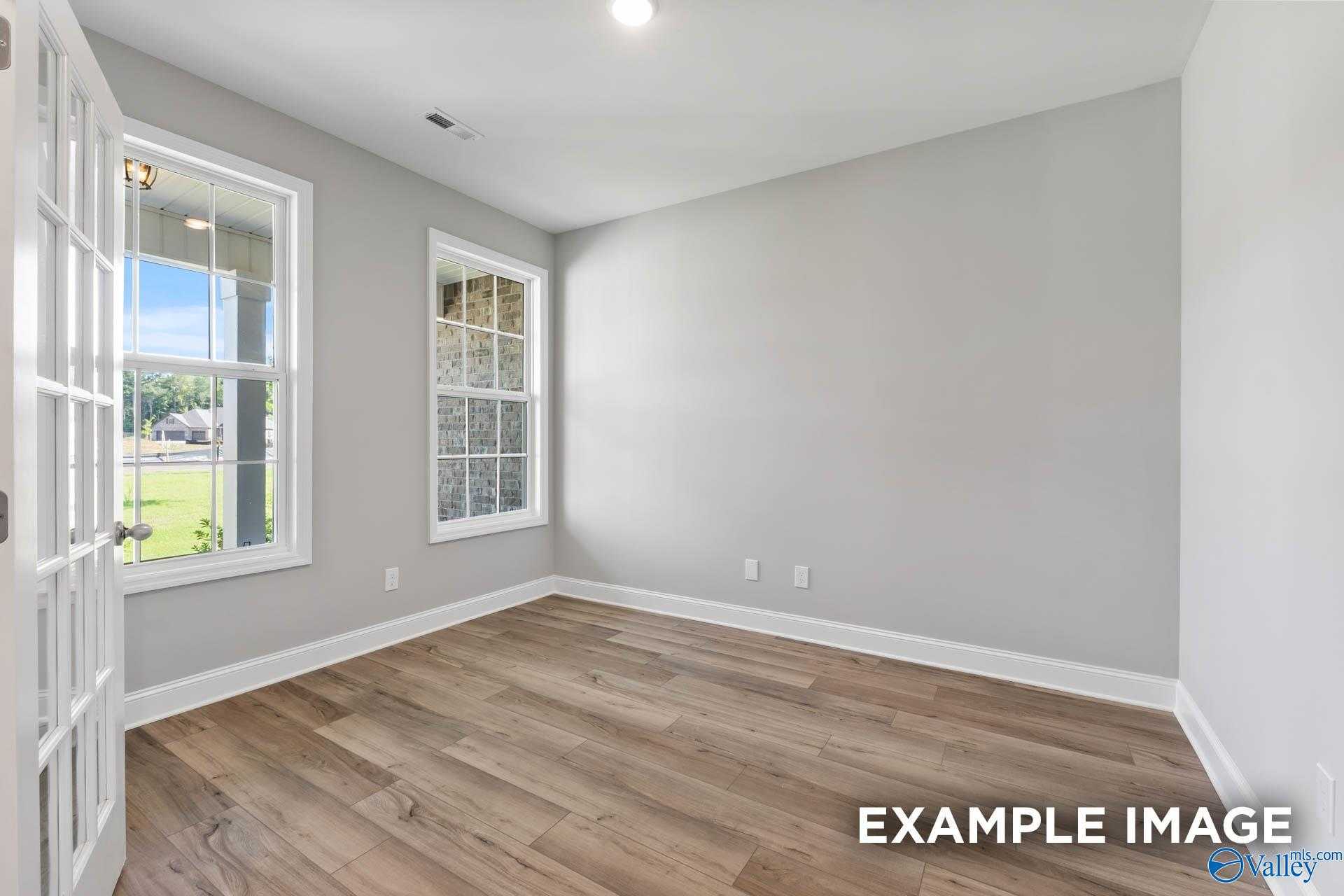 Bright bedroom with light gray walls, hardwood floors, and large windows overlooking green yard in Davidson Homes The Rockford D, Hazel Green, Alabama
