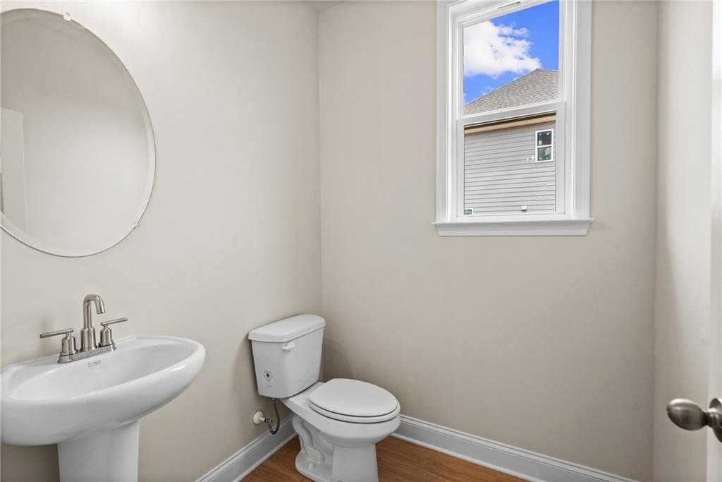 Bright powder room with round mirror, pedestal sink, white toilet, and window view in Davidson Homes The Hickory B at Wehunt Meadows, Hoschton