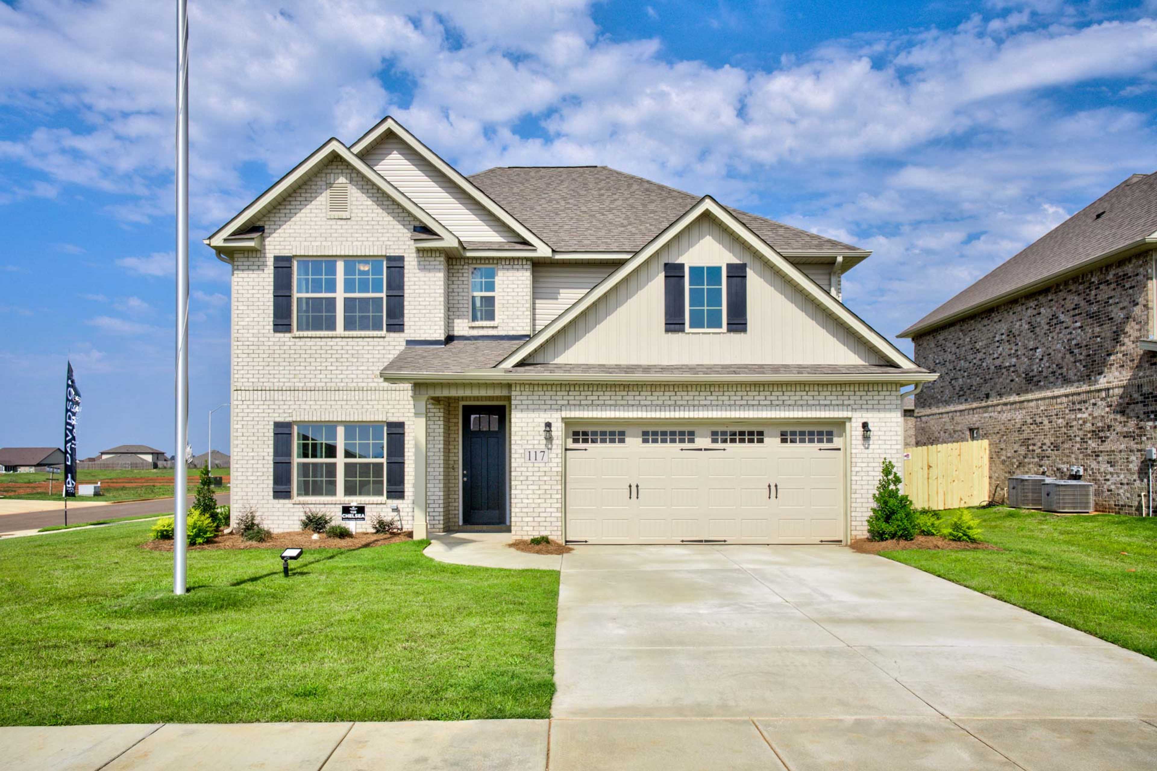 Two-story beige brick home exterior at Walker's Hill in Meridianville, Alabama with double garage, flagpole and lush green lawn