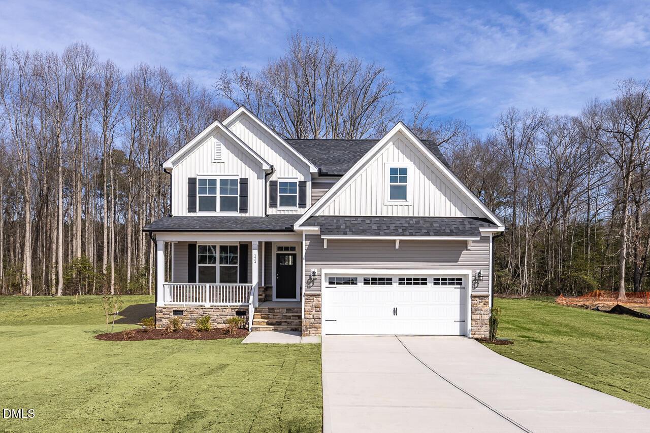 Modern two-story white farmhouse with black shutters, two-car garage, and wraparound porch amid wooded lot in Lillington, NC